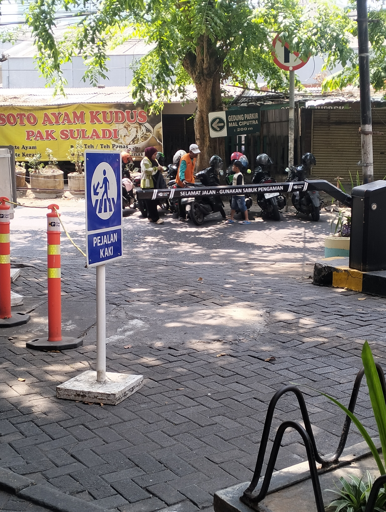 In Semarang, Indonesia, a street scene unfolds with a focus on pedestrian and vehicle access. A blue and white pedestrian crossing sign with an icon of a person walking is prominently displayed, accompanied by text in Indonesian that reads "PEJALAN KAKI," meaning "pedestrian." Adjacent to this sign, two orange traffic cones with yellow reflective bands are positioned on the paved sidewalk. Further down the street, a line of parked motorcycles is visible, some with helmets on their seats, suggesting recent use. Beyond the motorcycles, a black automatic gate barrier is installed, indicating a controlled entry point, possibly for a parking area. A sign with an arrow pointing left reads "GEDUNG PARKIR MAL CIPUTRA : 200m," translating to "Parking Building Ciputra Mall: 200m," confirming the location as near Ciputra Mall. The background features a building with a yellow banner advertising "SOTO AYAM KUDUS PAK SULADI," a popular Indonesian dish. The environment is characterized by a sunny day, with dappled sunlight filtering through the leaves of a large tree overhead, casting shadows on the patterned brick pavement. Several individuals are present, some standing near the motorcycles and others walking, contributing to the dynamic atmosphere of this urban setting.