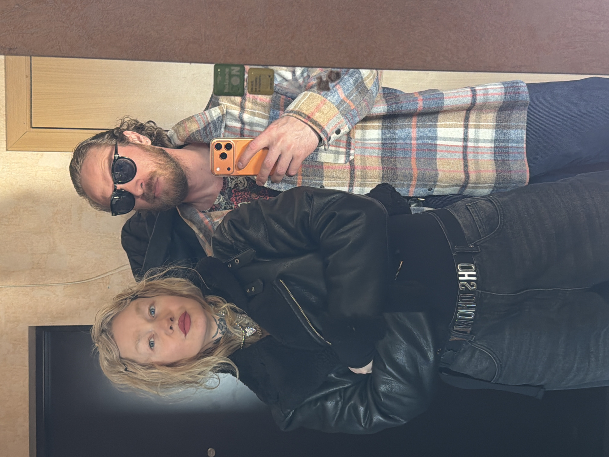 Two individuals are visible in the image, likely a couple, reflected in a mirror. The man, presumably holding the phone to take the picture, is wearing a plaid shirt and sunglasses. He has a beard and longer hair. The woman is wearing a black leather jacket. Her hair is blonde, and she has visible neck tattoos. They appear to be indoors, possibly in a fitting room or dressing area. The location is not identifiable as Saint Petersburg, Russia, based solely on the visual information provided. The background includes elements of furniture and a wall, offering limited clues about the specific location.
