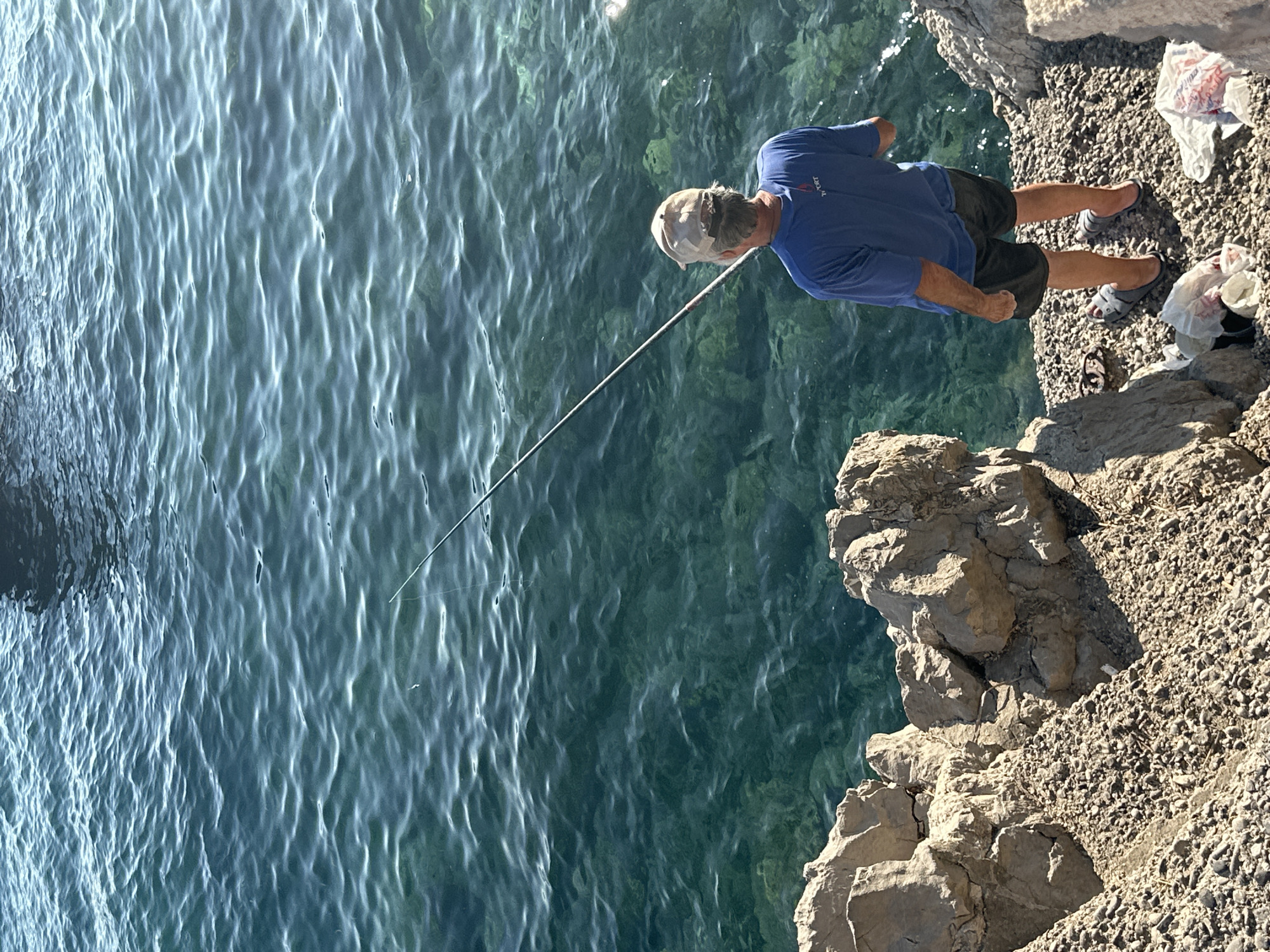 An individual stands on a rocky shoreline in Budva, Montenegro, casting a fishing line into the clear, turquoise water. The person, viewed from behind, wears a blue t-shirt, dark shorts, and a baseball cap. A fishing rod extends from their hand into the sea, with a thin line disappearing beneath the surface. The water reveals the rocky seabed beneath. The shore is composed of various sizes of stones and larger rock formations. In the upper right portion of the image, a dark silhouette of a boat is partially visible on the water. Two plastic bags, one containing white material, are on the ground near the individual's feet.