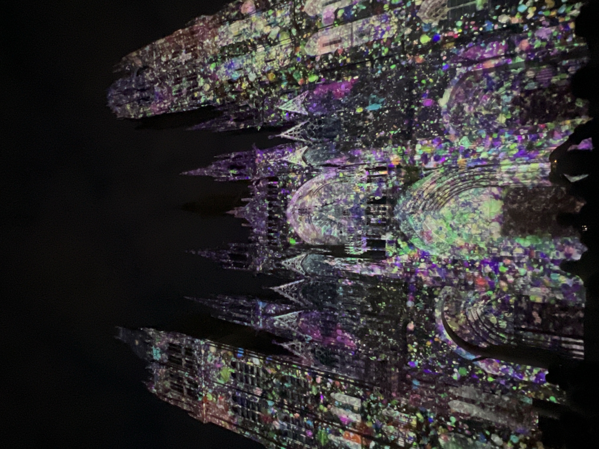 The image displays the facade of Rouen Cathedral (Cathédrale Notre-Dame de Rouen) in Rouen, France, at night. The Gothic architectural structure is illuminated by a dynamic, multi-colored light projection. The projection features numerous small, irregular shapes resembling paint splatters or confetti, primarily in shades of purple, green, blue, pink, and yellow, distributed across the entire surface of the cathedral. Two large, ornate towers rise on the left and right sides of the main facade, with intricate spires and arched elements visible in the central section. At the base of the image, several human silhouettes are discernible, indicating an audience viewing the projection. Some individuals appear to be holding up mobile devices, presumably to photograph or record the event. The sky above the cathedral is completely dark and featureless.