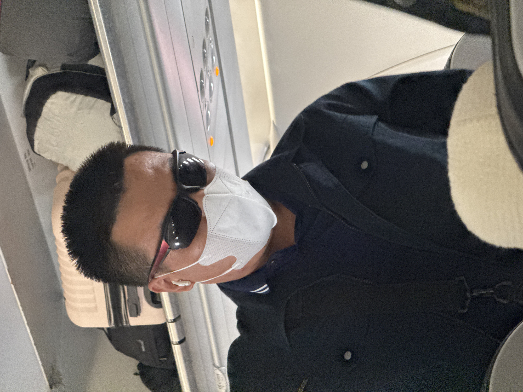 A man wearing sunglasses, a face mask, and a dark jacket is seated inside an airplane cabin. Luggage is visible in the overhead compartment. The lighting suggests it is daytime. There is no indication that this scene is in Hanoi, Vietnam.