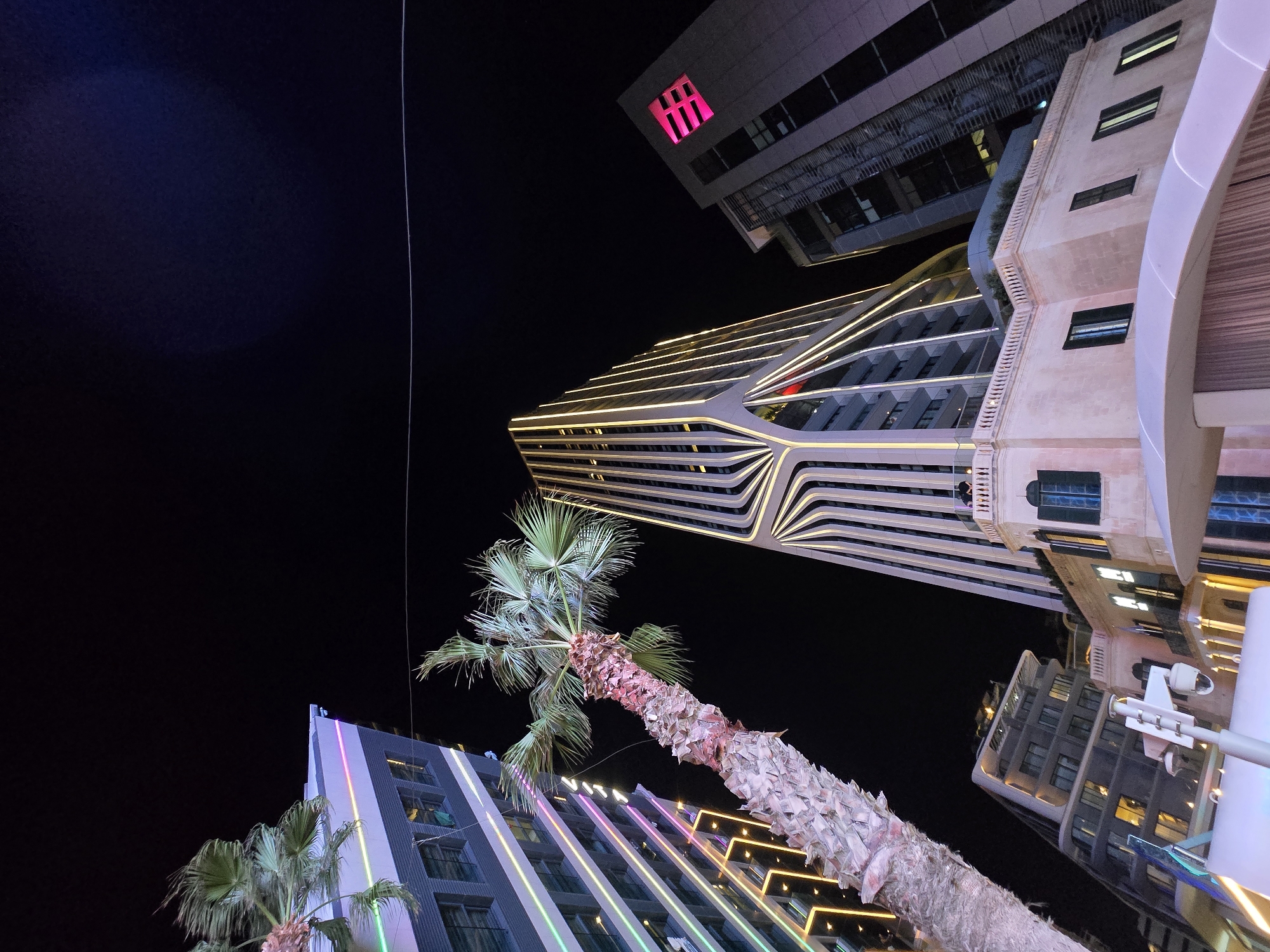 The image captures a night scene in Saint Julian's, Malta, focusing on modern architecture illuminated with colorful lights. In the foreground, a palm tree with a textured trunk and green fronds leans into the frame from the left. To the left of the palm tree is a tall, rectangular building with dark windows and vertical stripes of multi-colored LED lights, featuring pink, green, and yellow hues.
Dominating the center of the image is a striking, uniquely designed skyscraper. Its facade is composed of a series of curved, vertical white elements that create a flowing, almost liquid appearance. These elements are outlined with bright yellow LED lights, highlighting the building's distinctive shape against the dark night sky. A small section of red light is visible near the base of this building.
To the right of the central skyscraper is another tall building, this one more conventionally rectangular. Its facade is a darker gray, and it features a prominent, illuminated pink logo in the shape of a stylized 'E' within a square.
Below the central skyscraper and the building with the pink logo, a lower, more traditional building made of light-colored stone is visible. This building has arched windows and balconies, and it is also illuminated by interior lights, casting a warm glow. A white balcony railing runs along the top of this stone building.
In the lower-left quadrant, parts of a white structure with integrated lighting are present, along with a security camera mounted on a pole. The overall impression is one of a vibrant, modern urban environment at night, characterized by striking architectural designs and dynamic lighting.