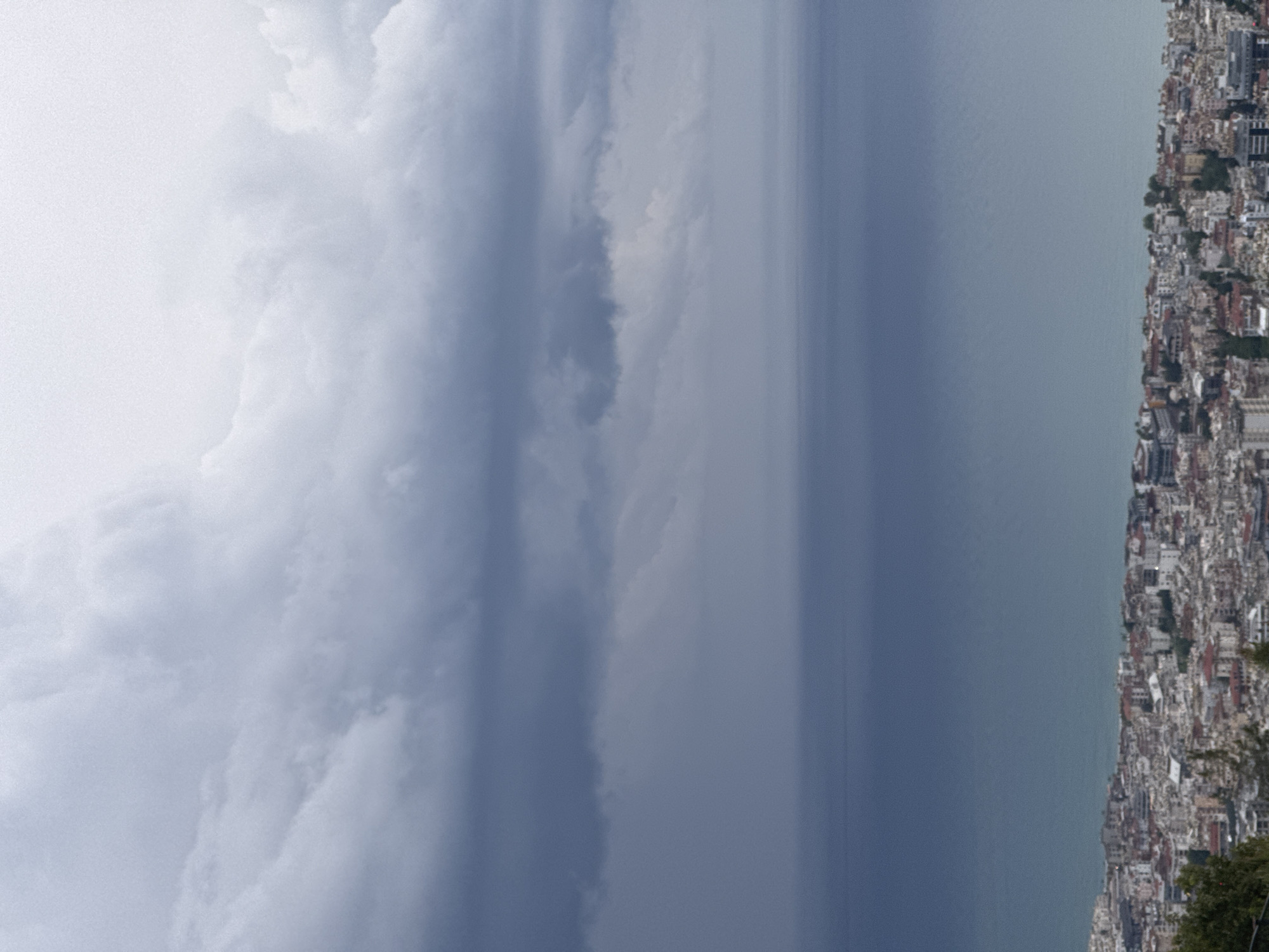The image shows a cityscape next to a body of water, with a cloudy sky above. The city appears densely built, with a variety of structures densely spaced together. The water is a uniform, light gray color. The sky is dominated by gray clouds. The location is Unknown, Unknown.