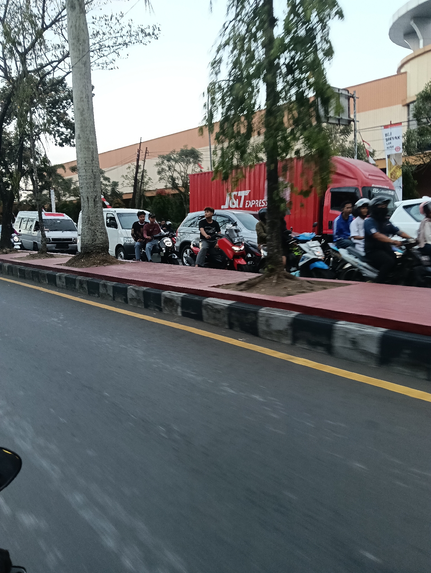 The image captures a street scene in Tasikmalaya, Indonesia, likely during daylight hours given the overall illumination. The foreground shows a stretch of asphalt road with a distinct yellow line marking a lane. The middle ground is dominated by a sidewalk area, paved with red and bordered by a black and white striped curb. A large tree with sparse leaves stands prominently on this sidewalk, near a light pole.
To the right of the sidewalk, several vehicles are visible. A red shipping container with "J&T EXPRESS" branding is parked, partially obscuring some of the scene. In front of the container, there are multiple motorbikes. Three young men are seated on two of these motorbikes, with one man appearing to be talking to another. Another motorbike is occupied by a person in a black helmet, facing forward, suggesting movement or readiness to move. Further to the right, a white van is parked. Another motorbike with a rider and passenger is in motion, captured with motion blur. In the background, a large building with a tan and orange facade is visible, with a curved roof structure at the top. A banner with Indonesian text is also present on the building. The sky is overcast and appears bright. The overall atmosphere suggests a typical urban street with active transportation.