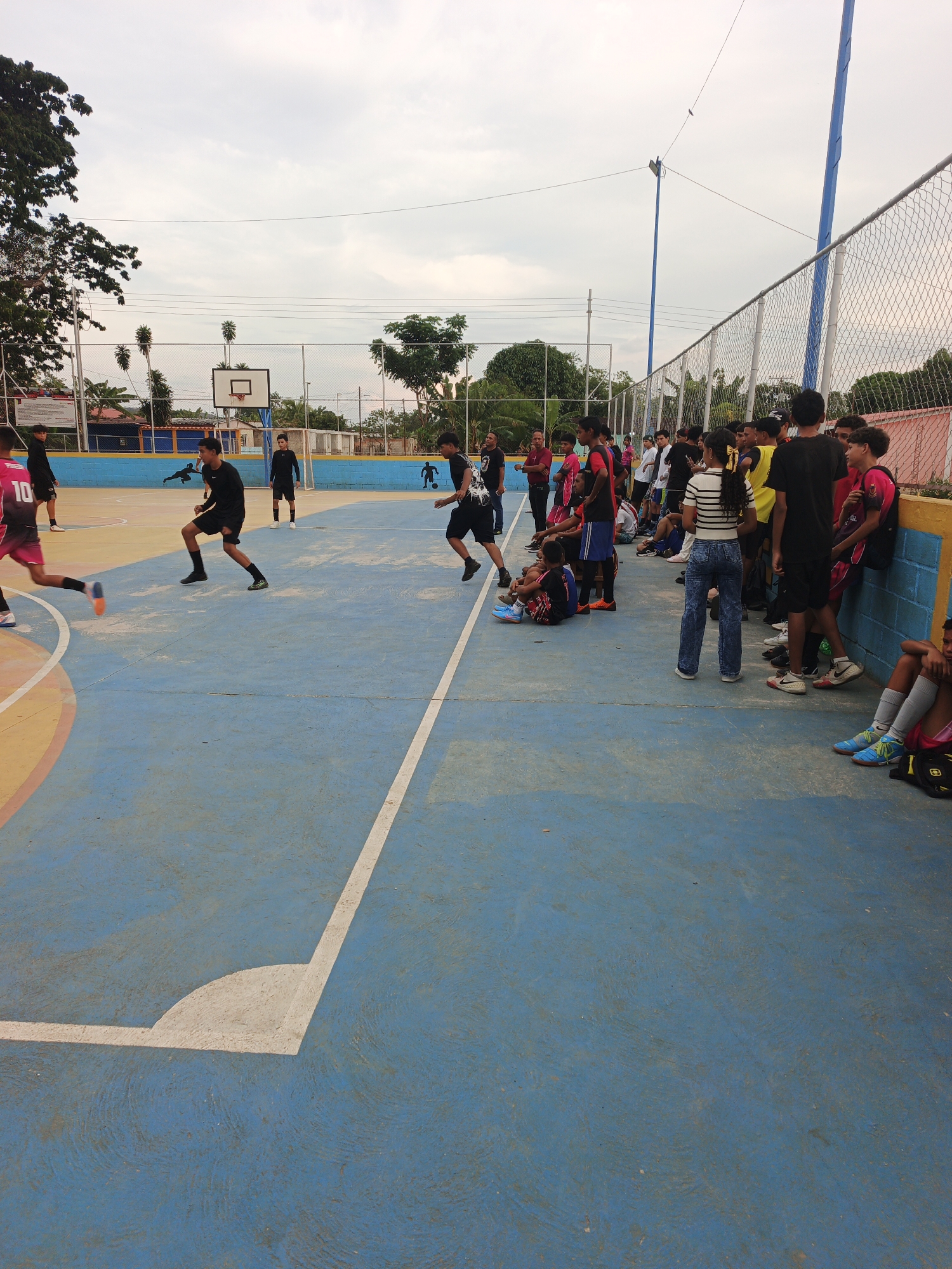 This image depicts an active outdoor futsal court scene in Tinaquillo, Venezuela, during what appears to be late afternoon or early evening under an overcast sky. The court itself is primarily blue with yellow and white markings, including a clear corner arc in the foreground.
Numerous young people, predominantly male teenagers and young adults, are present. A futsal or soccer game is underway, with several players actively engaged on the court. One player in a red jersey with the number "10" is in motion on the left, while others in dark attire are also participating. In the background, a white soccer goal and a basketball hoop are visible, indicating a multi-sport facility.
Along the right side of the court, a larger group of young spectators and resting players gathers. Many are standing or sitting on a low blue and yellow concrete wall, observing the game. A girl in a striped shirt and blue jeans stands among them. The general mood seems relaxed and social, typical of a community recreational event.
The court is surrounded by a chain-link fence supported by tall blue poles. Beyond the fence, various trees and modest buildings are visible, adding to the community setting. While no specific text is fully legible other than the "10" on the jersey and some faint brand logos on shoes, the scene clearly conveys an energetic and informal sports gathering.