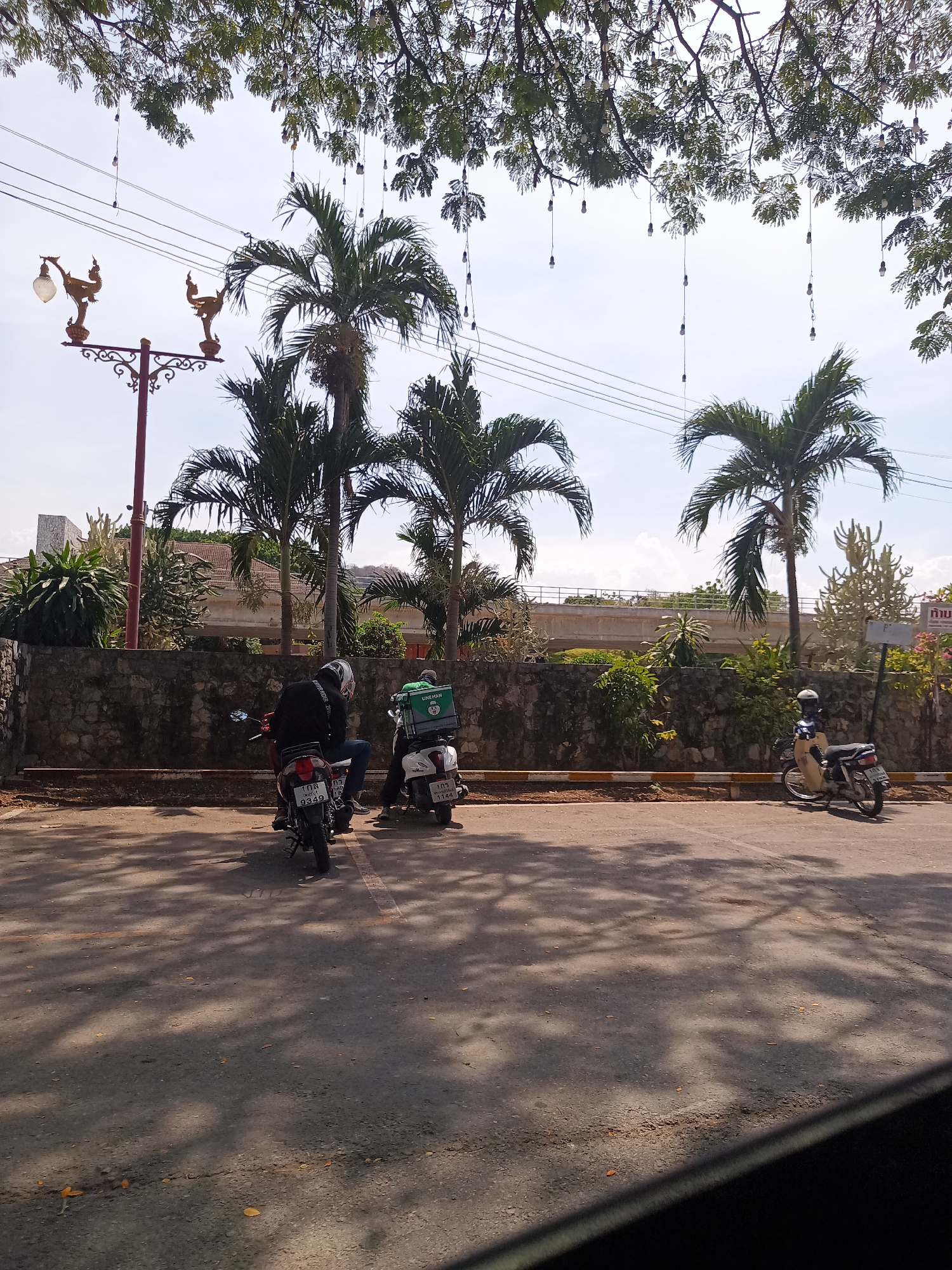 The photograph depicts a street scene in Hua Hin City Municipality, Thailand, with palm trees and overhead string lights. Two motorcycles are parked along the curb with their drivers in close proximity, while another is seen farther away. The street has visible shadows, indicating sunlight. The background features a stone wall with vegetation and a raised structure in the distance. A decorative lamppost is on the left.