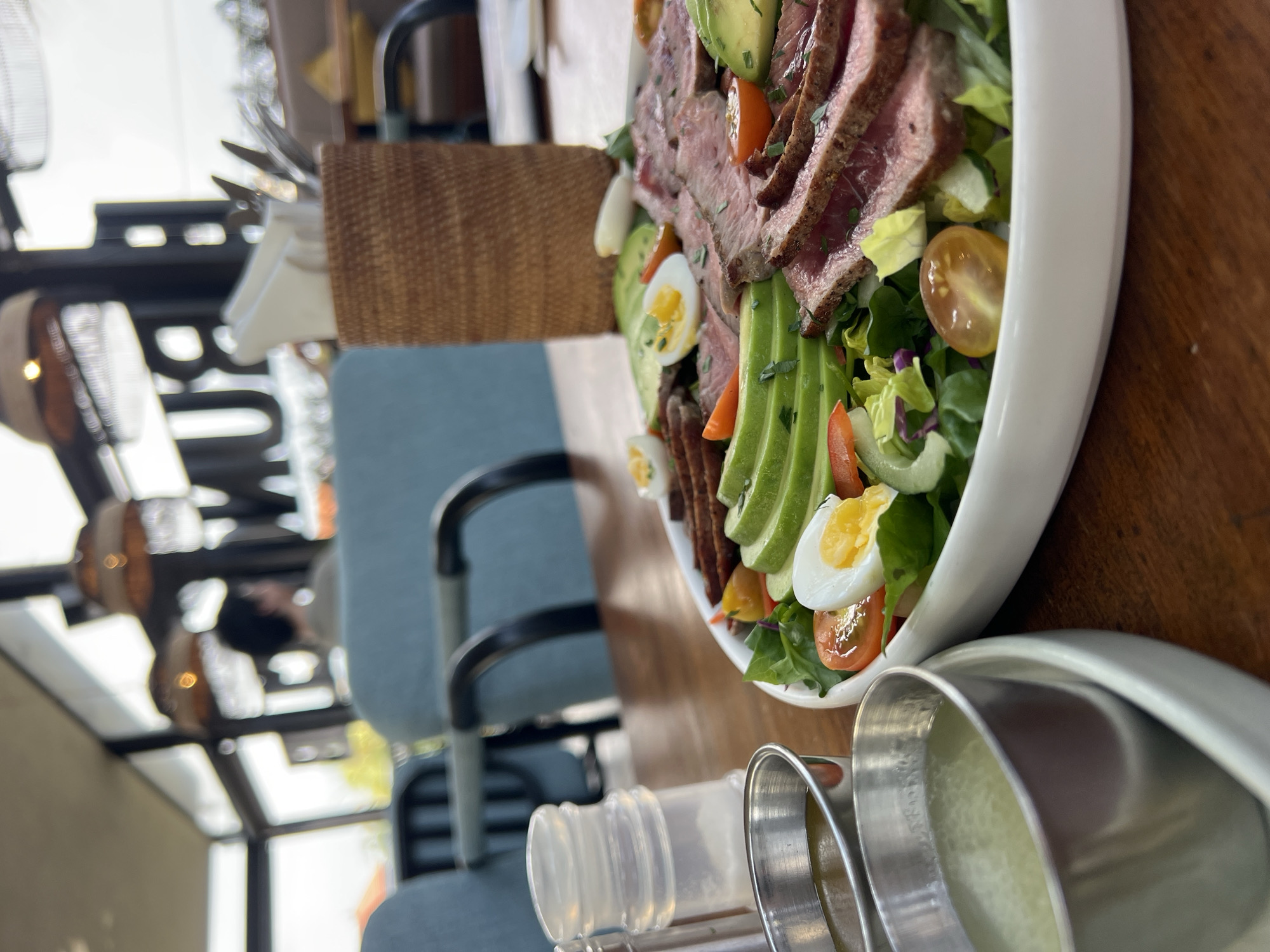 A close-up, eye-level shot captures a substantial steak salad presented on a white plate, situated on a wooden table. The salad is a vibrant medley of ingredients, featuring thinly sliced, medium-rare steak arranged artfully over a bed of mixed greens. Slices of ripe avocado fan out across the plate, interspersed with halved cherry tomatoes, sliced hard-boiled eggs, and slivers of cucumber and red bell pepper. The dish is garnished with finely chopped fresh herbs. To the left of the salad, two small, metallic condiment cups hold dressings, one a lighter, possibly vinaigrette, and the other a darker liquid. A clear plastic salt shaker is also visible.
The background of the image reveals the interior of a restaurant, likely in Mengwi, Indonesia. Softly lit by ambient light from decorative pendant lamps with woven shades, the setting appears casual yet stylish. In the mid-ground, two light blue chairs with dark metal frames are partially visible. Further back, through a large window, a blurry scene of outdoor greenery and distant structures can be discerned. A person with dark hair is seated in one of the chairs, their back mostly to the camera. The restaurant's interior features large windows, offering glimpses of the outside environment and an illuminated sign with the word "BUN" is visible in the distance. A woven basket, possibly holding napkins and cutlery, stands on the table behind the salad. The overall atmosphere suggests a meal being enjoyed in a well-lit, contemporary dining establishment.