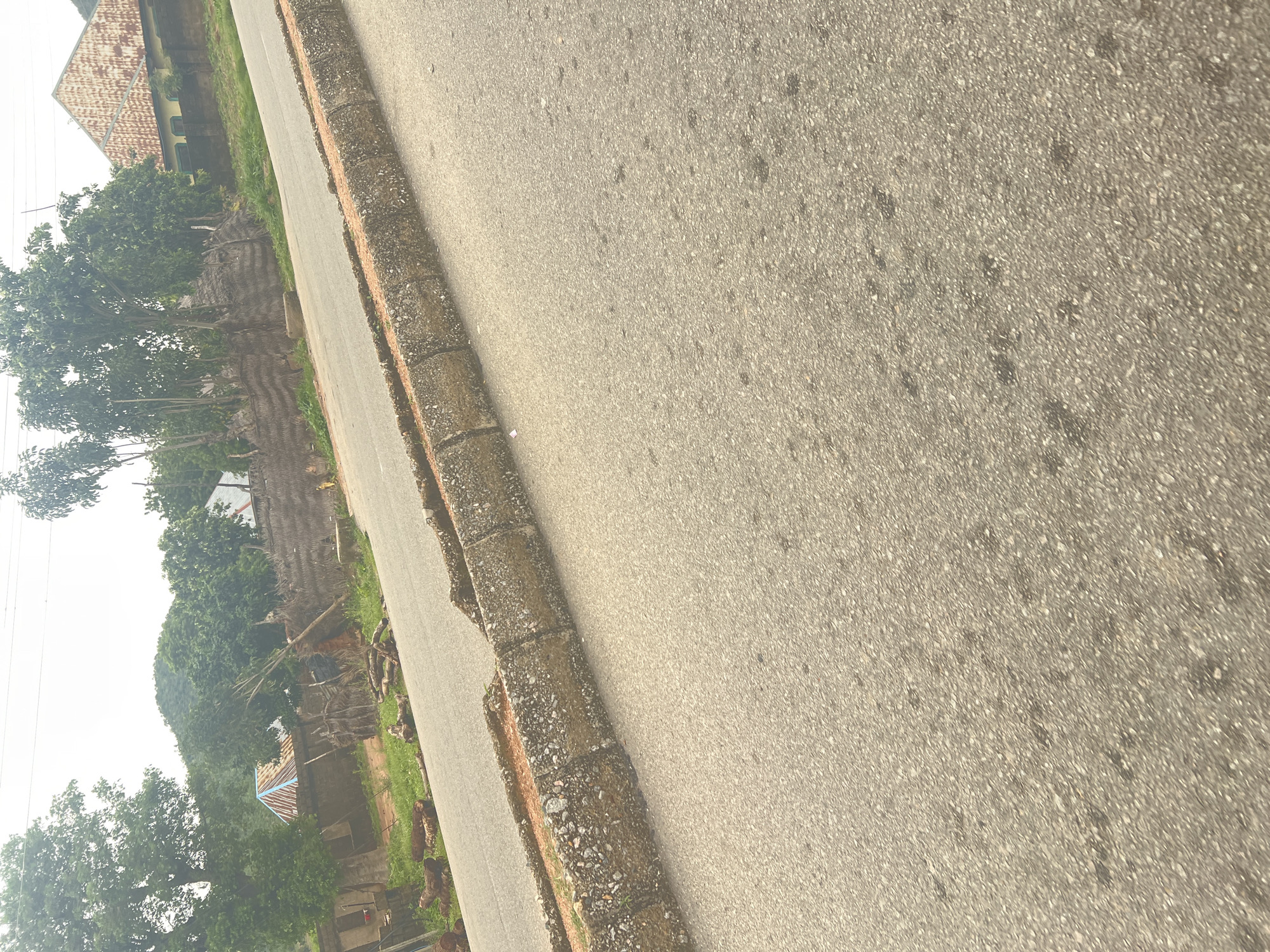 The image shows a road in Kona Garu, Nigeria. The road is paved and has a curb made of concrete blocks. On the left side of the road, there are some houses with thatched roofs and corrugated iron roofs. There are also some trees and vegetation. On the right side of the road, there is a house with a corrugated iron roof and a wall. The sky is overcast. The image appears to have been taken from a low angle, looking across the road.