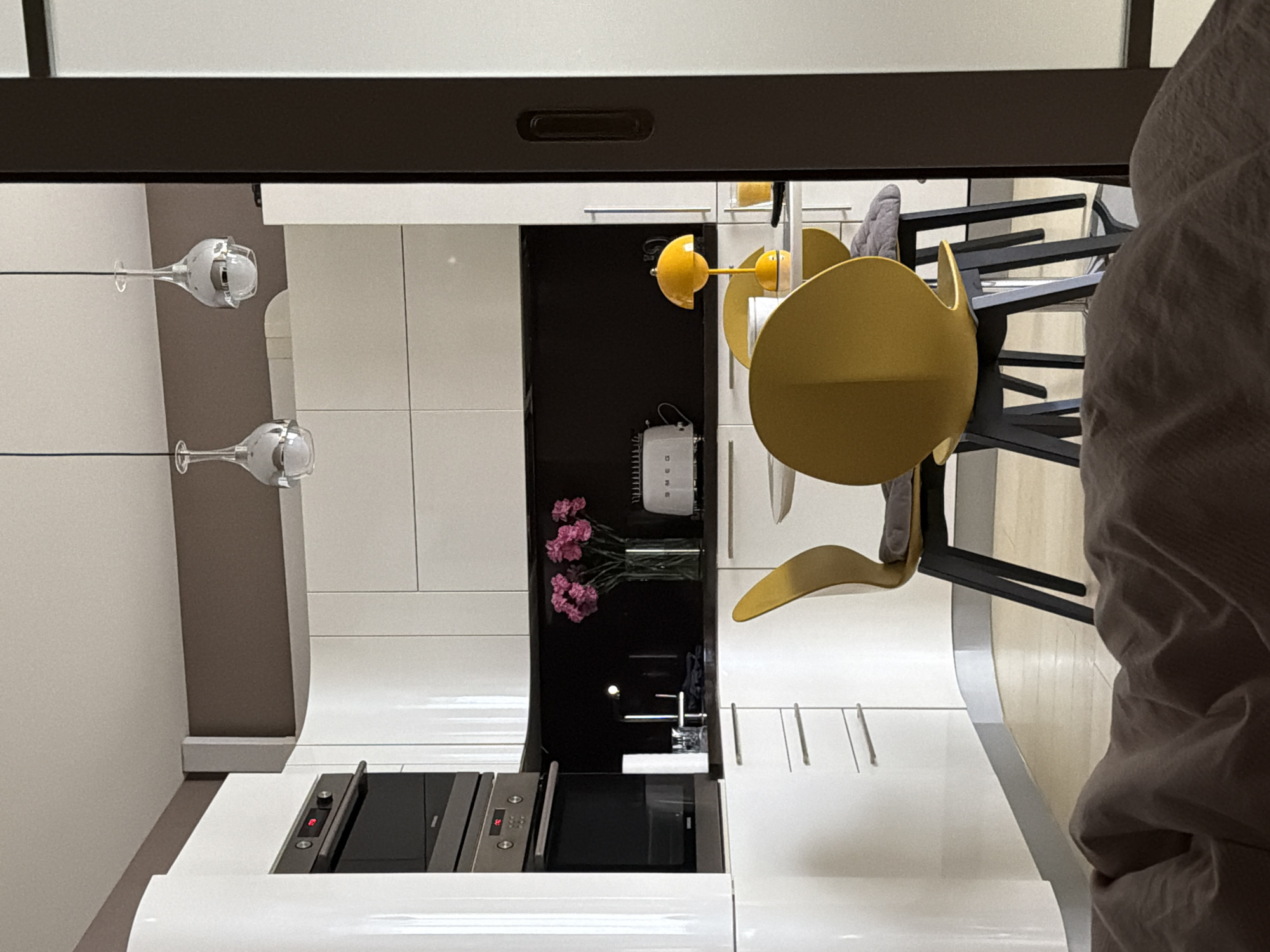 The image shows an interior, likely a kitchen, in Khimki, Russia. The kitchen features white cabinets, a built-in oven and stovetop, and a dark-colored niche housing a vase of pink flowers, a Smeg appliance, and a sink with a faucet. A yellow-colored lamp with a circular shade stands on the countertop near a stack of black chairs. Two glass pendant lights are visible against a light-colored wall. A person wearing a light-colored shirt is partially visible on the right side of the image.