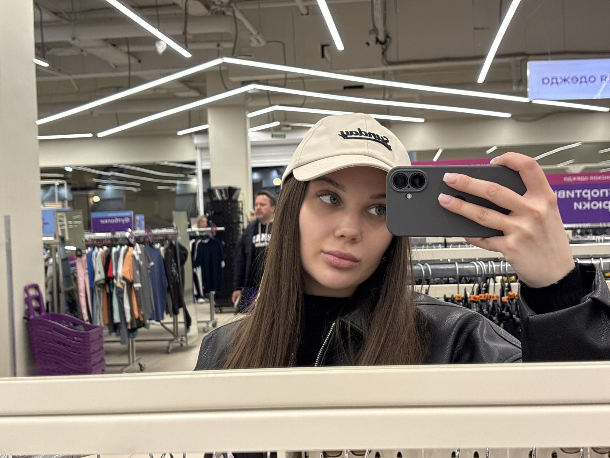 A young woman is captured in a mirror selfie inside what appears to be a retail store, likely in Yekaterinburg, Russia. She holds a dark-colored smartphone with two visible camera lenses in her right hand, obscuring part of her face. She wears a light beige baseball cap with a dark, stylized, mirrored "Umbro" logo, a black turtleneck, and a black faux leather jacket. Her long, dark brown hair falls over her shoulders, and her gaze is directed away from the phone camera, slightly to her right.
The background, reflected in the mirror, reveals an indoor commercial space with bright, linear fluorescent lighting strips on the ceiling. Numerous clothing racks with various garments are visible throughout the store. In the mid-ground, a man with short brown hair and a dark jacket is reflected. To the left, a stack of purple shopping baskets is positioned near a clothing rack.
Visible text in the reflections includes a sign in the upper right that reads "ОДЖЕДО R" which, when un-mirrored, translates to "ОДЕЖДА R" (Clothing R) in Cyrillic. Below this, there's a blurry reflection of another sign, with faintly discernible Cyrillic text that appears to be "ОДЕЖДА НА КАЖДЫЙ ДЕНЬ" (Clothing for Every Day). The scene suggests the woman is shopping or trying on clothes. The lighting indicates it is likely during business hours, either day or evening.