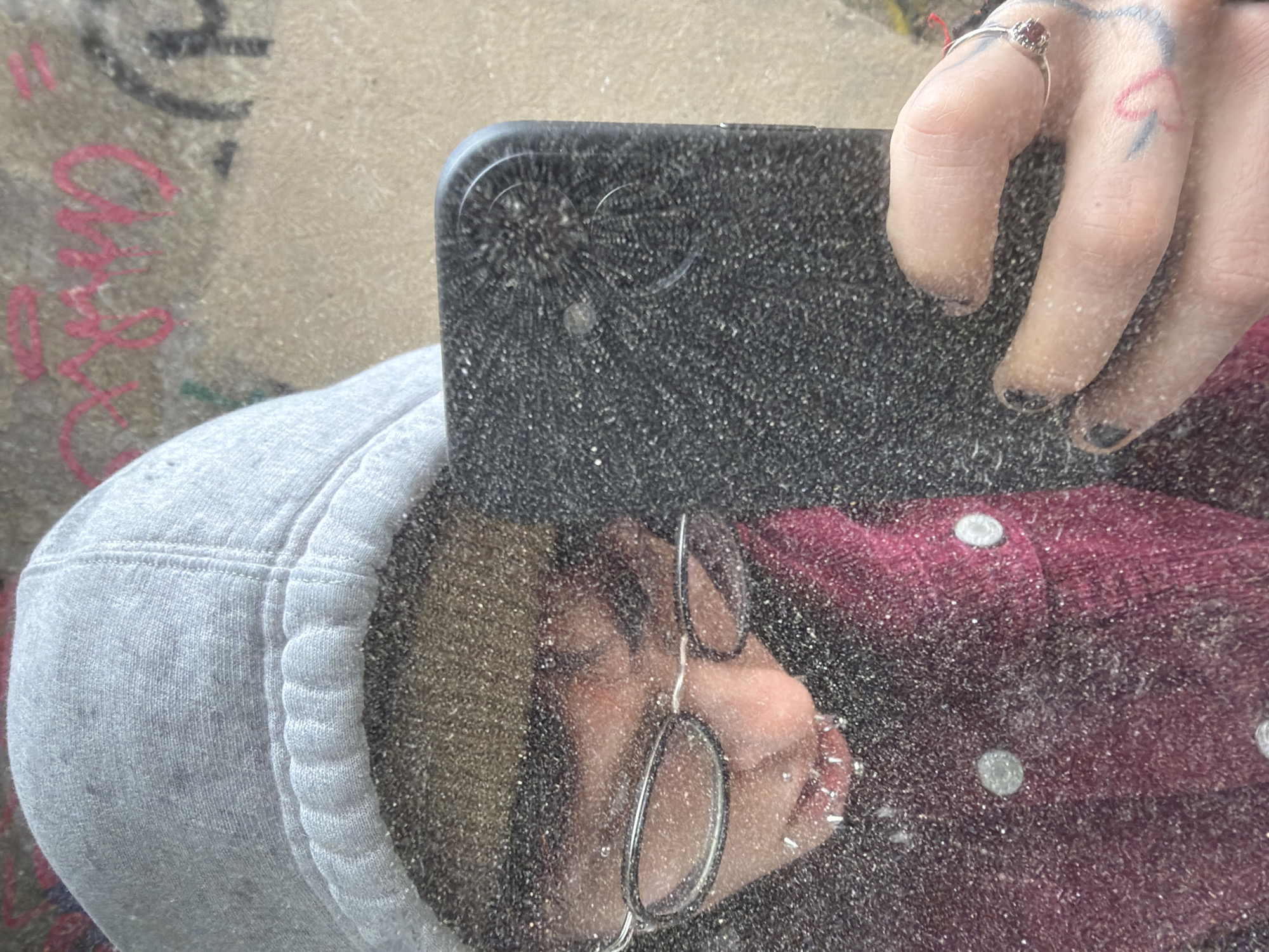 A close-up, slightly tilted, self-portrait shot is taken of a person reflecting in a dusty mirror. The person is wearing a grey hoodie and a maroon button-down shirt, partially visible with its white buttons. They are wearing round-framed glasses. Their fingers, adorned with a ring and a heart tattoo, are holding up a black smartphone with a cracked camera lens. A concrete surface with graffiti is visible in the background, suggesting an urban setting. Insufficient data to determine the specific location in Sofia, Bulgaria.