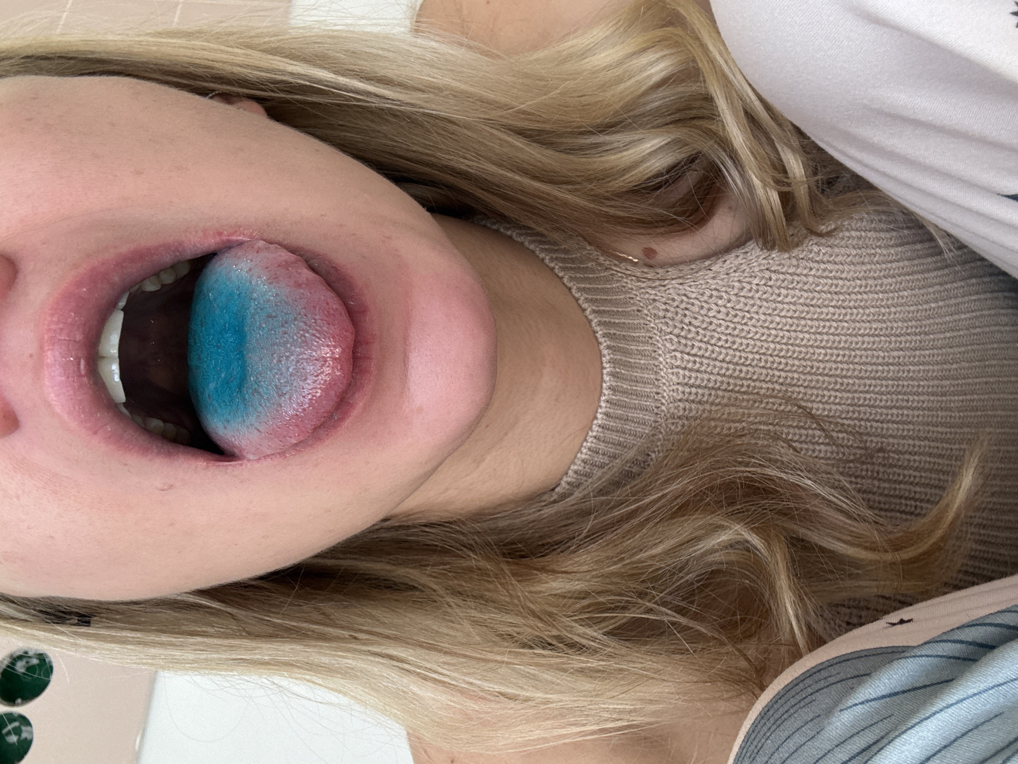 A close-up, rotated image shows a fair-skinned person with blonde hair. The person is wearing a tan-colored knit shirt. Their mouth is open, revealing white teeth and a tongue that is blue towards the tip and pink towards the base. The image suggests the person has consumed something that has stained their tongue blue. The location is unknown.