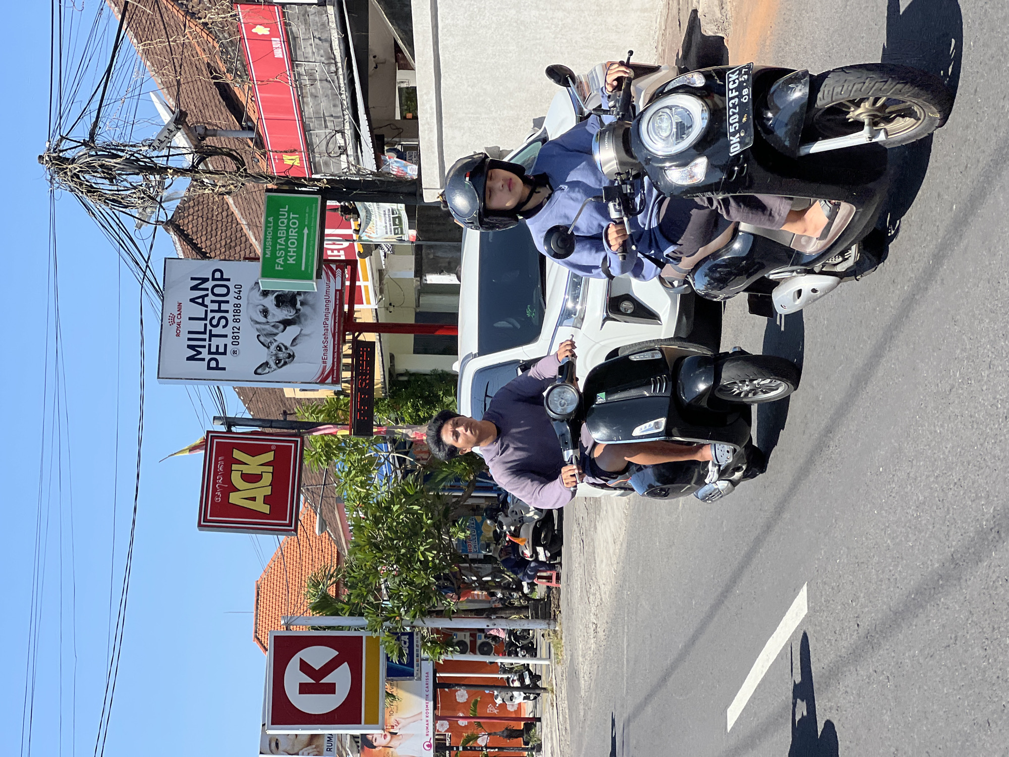 The image captures a street scene in Denpasar, Indonesia, under a bright, clear sky. Two individuals are riding scooters down a paved road. The person in the foreground, wearing a blue long-sleeved shirt and a black helmet, rides a black Honda scooter with a license plate reading "DK 5023 FCK". To their left and slightly behind, another person, wearing a purple long-sleeved shirt and dark shorts, rides a black scooter. A white car is visible behind them. Several signs are present along the roadside, including one for "MILLAN PETSHOP", a red sign with "ACK" in yellow lettering, and a red and white sign with a "K" logo. Lush green trees line the street, and buildings with tiled roofs are visible in the background. The environment suggests a typical urban setting in Indonesia, possibly a commercial area with advertisements.