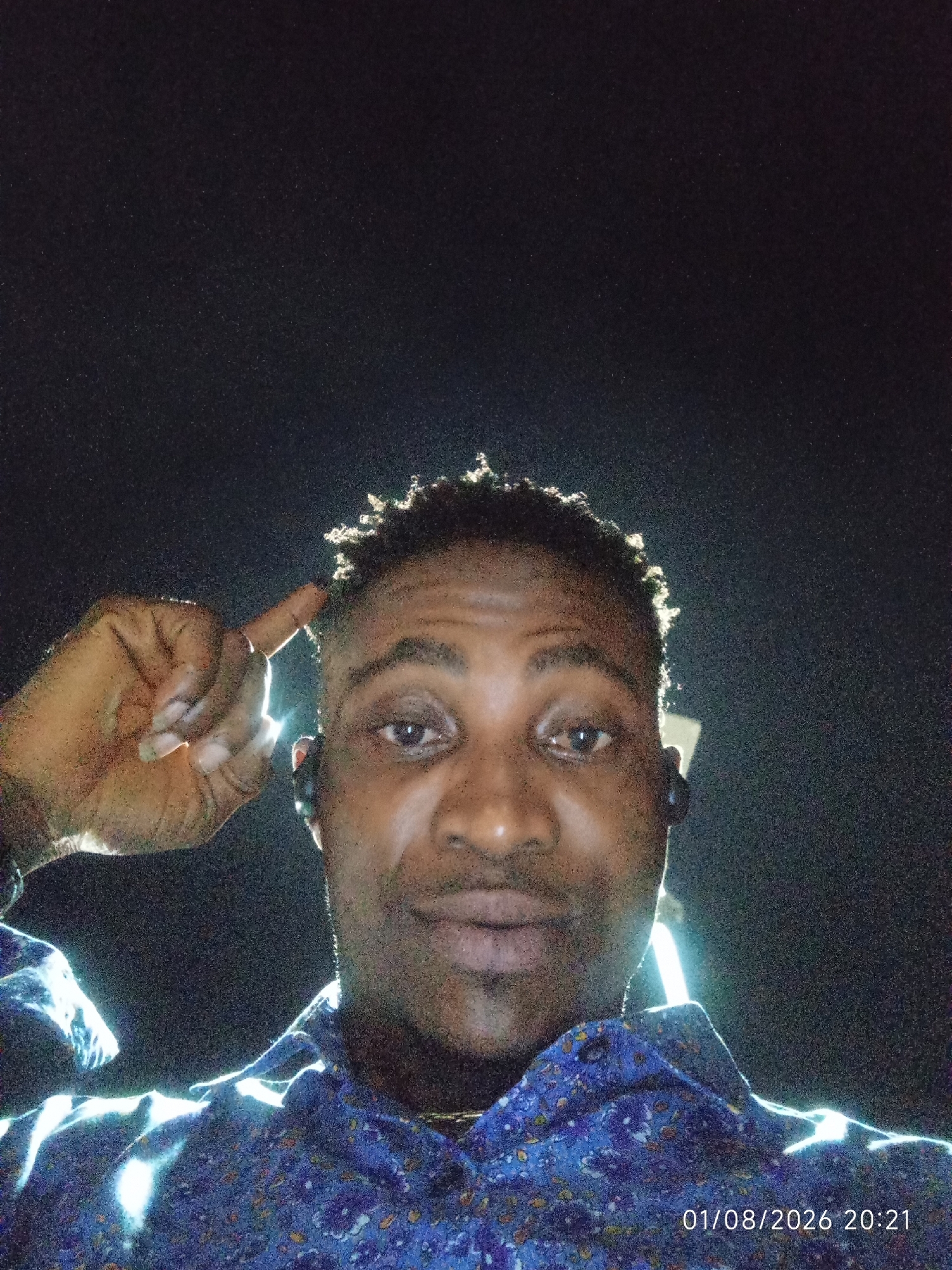 A man with dark skin, short, curly hair, and a patterned blue shirt stands in front of a dark background. He is looking towards the camera and has his right index finger pointing towards his forehead. The lighting is strong and appears to be coming from below and to the right, creating bright highlights on his face and shirt. A timestamp in the lower right corner indicates the image was taken on 01/08/2026 at 20:21. The background is uniformly dark, suggesting a nighttime setting or a dimly lit interior. There are no other individuals or objects visible in the frame. The specific location in Adankolo, Nigeria, cannot be determined from the image alone.