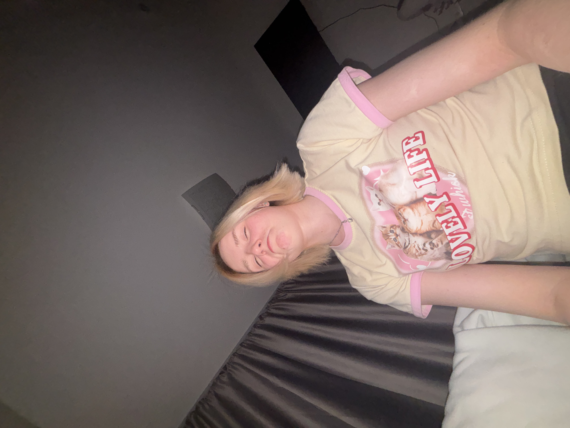A blonde-haired person is lying on a bed. The person is wearing a pale yellow t-shirt with pink trim and a graphic of cats. The bed has a gray duvet and a white pillow. The background is a gray wall. A black object is partially visible in the upper portion of the frame. It is not possible to determine the location as Vsevolozhsk, Russia from the image.