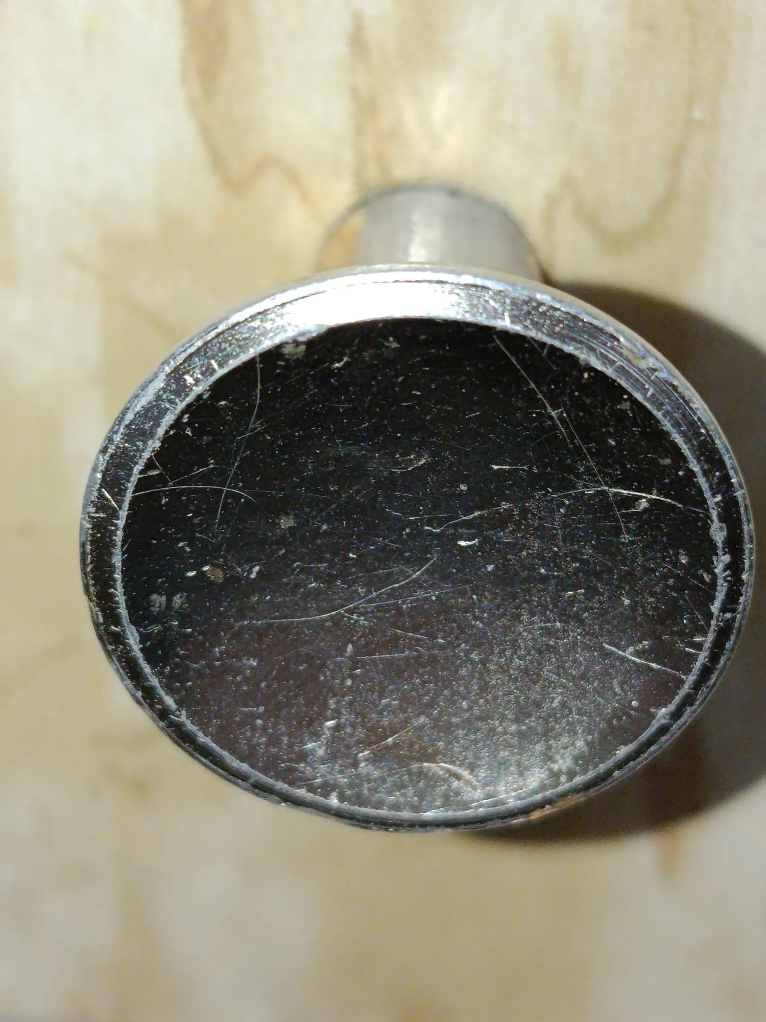 The image shows a close-up, top-down view of a metal object with a dark, scratched interior, possibly a camera lens cap or a similar cylindrical object. The exterior appears to be made of brushed metal. The background is a light-colored, textured surface, resembling wood grain. The lighting suggests an indoor environment, with a shadow cast by the object on the background to the right. There are no people or other identifiable objects in the scene. The location in Tashkent, Uzbekistan, is not discernible from the visual information provided in the image.