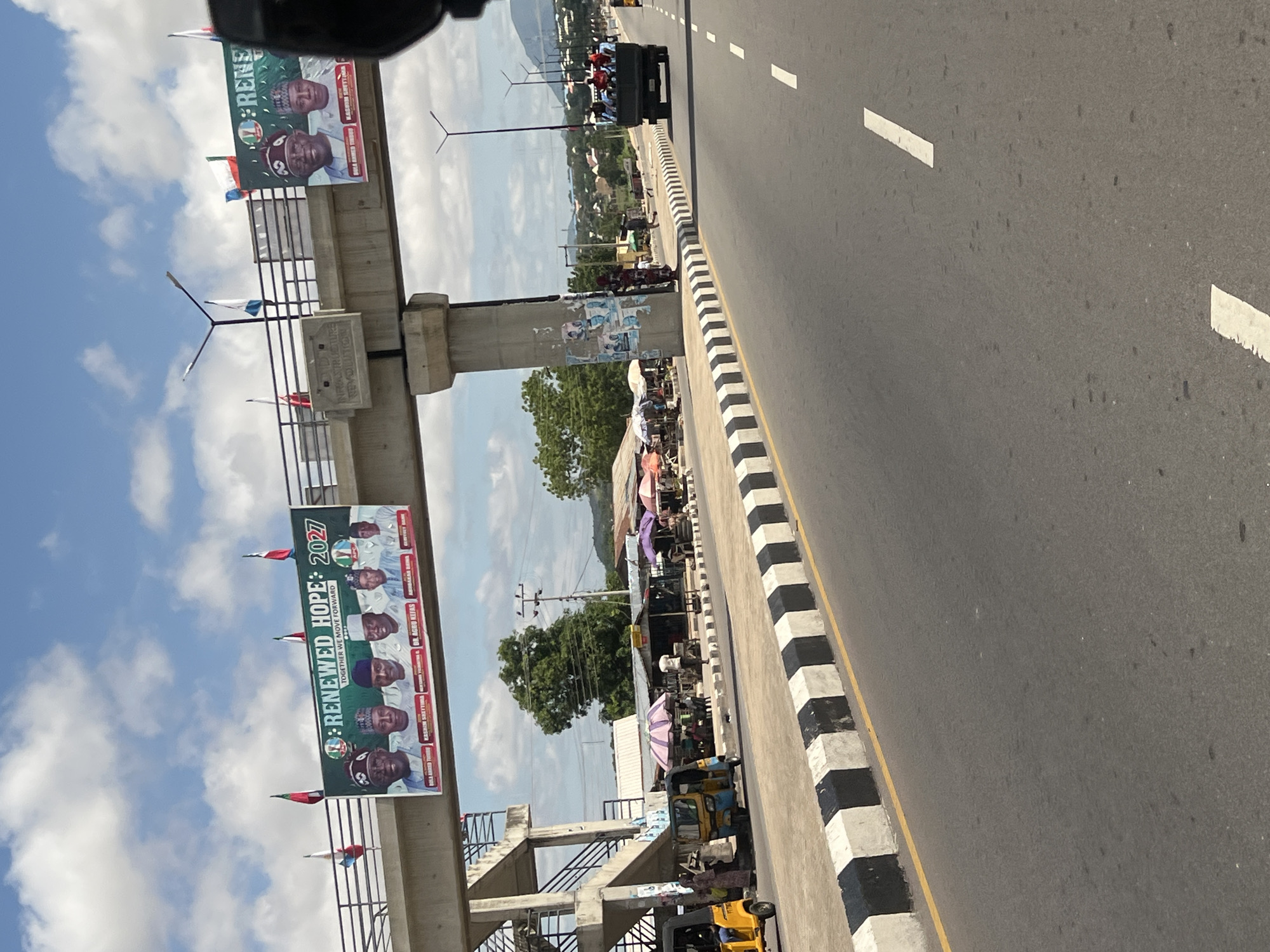 This image depicts a bustling urban scene in Jalingo, Nigeria, centered around a multi-lane asphalt road and an overhead pedestrian overpass, viewed under a partly cloudy sky.
The prominent feature is a large green banner stretched across the concrete pedestrian overpass. The banner displays "RENEWED HOPE : 2027" and "TOGETHER WE MOVE FORWARD" with the logo of the All Progressives Congress (APC) political party. It features headshots of six men in traditional attire; identifiable names below them include "BOLA AHMED TINUBU", "KASHIM SHETTIMA", "DR. AGWU KIFAS", "ABUBAKAR BARAU", and "GUMNEY DANE". Small Nigerian flags and other colored flags are affixed to the overpass's metal railing. A concrete block on the overpass structure bears the inscription "INFRASTRUCTURE REVOLUTION".
Below the overpass, the road features white dashed lane markings and is bordered by a black and white striped curb with a thin yellow line. To the left of the road, a vibrant street market operates along a paved shoulder. Numerous vendors are set up under colorful umbrellas (pink, purple, green, white) and makeshift stalls. Several people, dressed in a mix of traditional and casual attire, are gathered around these stalls. Two yellow three-wheeled motorized rickshaws (keke napep) are parked near the market area, with individuals standing by them.
Further down the road on the right, a dark green utility truck is visible, with its open bed filled with numerous passengers, many wearing red tops. The truck is moving away from the viewer. In the background, beyond the market and residential structures with corrugated roofs, distant hills are discernible. Multiple streetlights with black poles and multi-arm fixtures are positioned along the roadside. The image appears to be taken from a vehicle, with a portion of the vehicle's interior visible on the far right foreground.