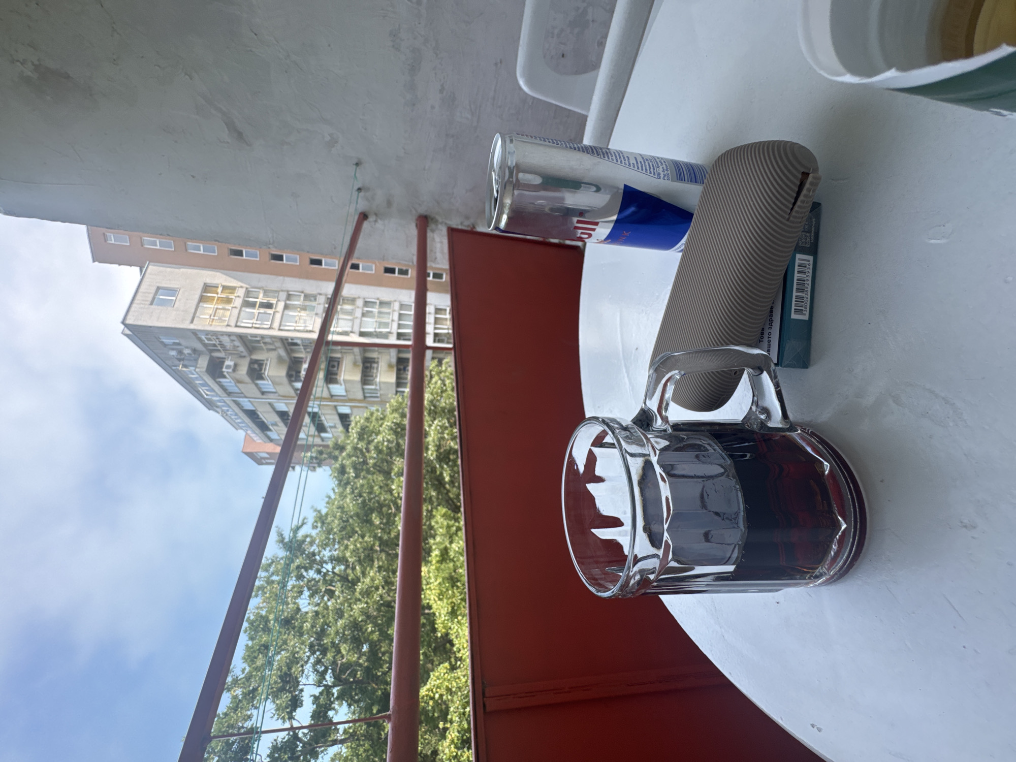 This image captures a bright, daytime scene on what appears to be a balcony or outdoor patio. A white, round table in the foreground holds a clear glass mug with a dark liquid, a silver Red Bull can, and a textured grey object, possibly a vape device, with a small broken section. A rectangular green and blue package is partially visible beneath the grey object, displaying a barcode and Cyrillic text including "Toba" and "здраве" (health).
Beyond the table, a red metal railing marks the edge of the space. Behind the railing, dense green trees fill the midground, suggesting a natural element in an urban setting. In the background, several multi-story buildings with numerous windows are visible under a clear blue sky.
The objects on the table suggest a casual, relaxed moment, possibly enjoying a drink and a vape. The time of day is sunny and clear, indicating fair weather. The Cyrillic text on the package provides a strong clue to the geographical location, likely a Slavic-speaking country. No people are present in the scene.