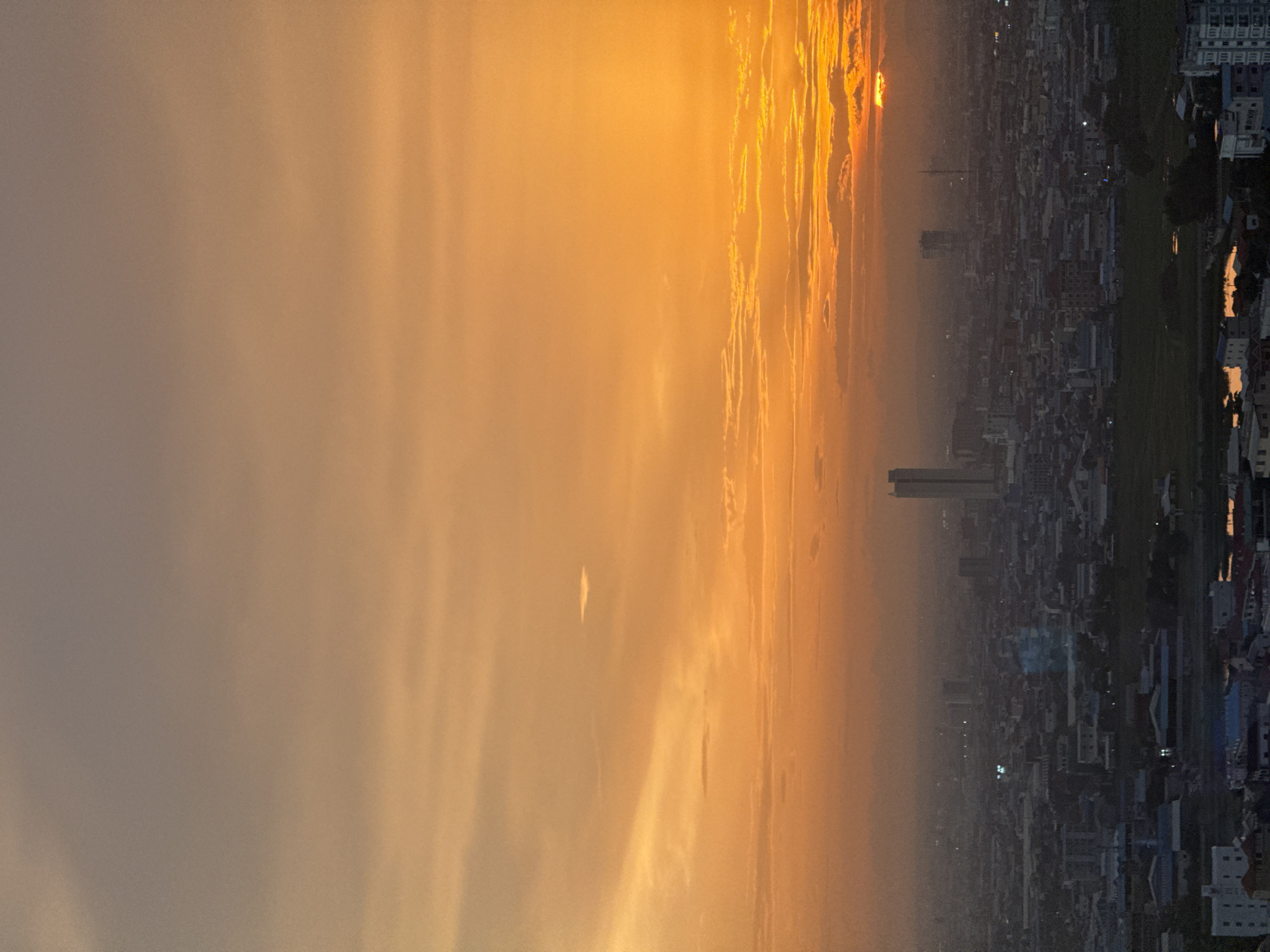 A cityscape is viewed from a high vantage point at sunset. The sky is a gradient of orange and yellow, with thin clouds streaking across. The sun is low on the horizon, partially obscured by clouds, casting a warm glow over the scene. Below, the city stretches out, a dense collection of buildings of varying heights. Several tall skyscrapers are visible, including one prominent, slender building towards the right of the frame. The urban landscape appears somewhat hazy, suggesting atmospheric conditions or a light dust. Some pockets of green space, possibly parks or undeveloped land, are interspersed within the urban fabric. The foreground shows a closer view of rooftops and some structures, indicating the perspective is from a building within or overlooking the city. The general impression is of a sprawling urban environment at the close of day.