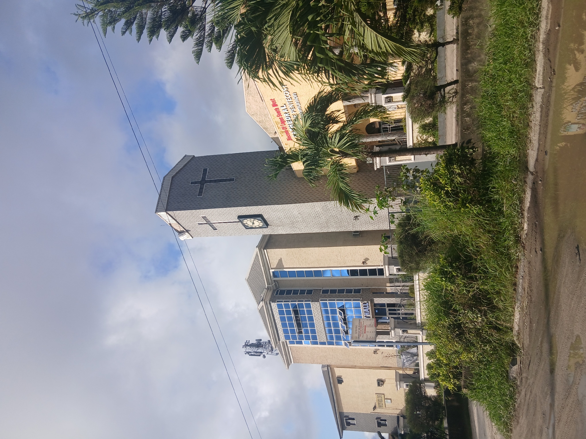 A wide shot captures the exterior of a church in Satellite Town, Nigeria, under a partly cloudy sky. The foreground is dominated by a muddy, waterlogged dirt road reflecting the surrounding environment. Lush green vegetation, including tall grasses and bushes, lines the edge of the road and the church property.
The main subject is a multi-story church building. The central structure is a tall, gray tower with a clock face and a prominent black cross. Adjacent to the tower is a lighter-colored building with multiple floors and large blue-tinted windows. A sign above the entrance reads "CATHEDRAL CHURCH OF THE PENTECOST" and "HALL." Another building, also lighter in color, is visible to the left. The church appears to be an active place of worship, indicated by its signage and architectural style. The surrounding environment suggests a tropical or sub-tropical climate, given the vegetation. No people are visible in the frame.