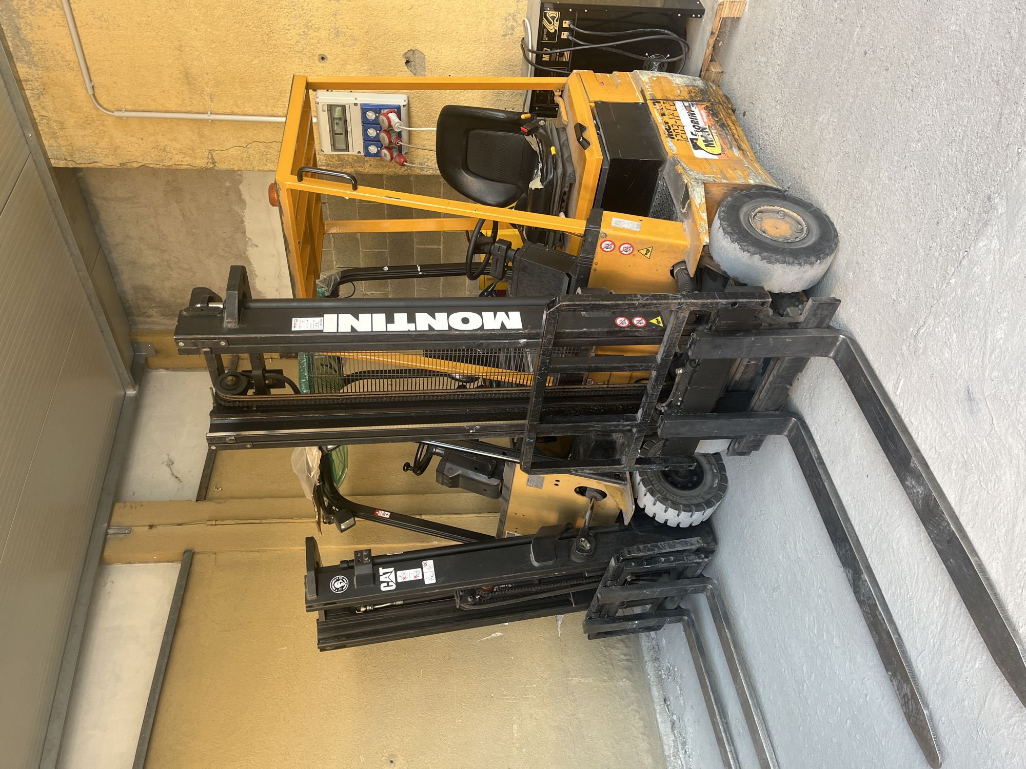 Two forklift trucks are parked next to each other in an outdoor, covered area with a concrete floor and yellow and grey walls. The forklift on the right is yellow and black and has the brand "MONTINI" visible on its mast. It has a black seat and a control panel with electrical outlets attached to its frame. The forklift on the left is black and has the "CAT" logo visible on its mast. Both forklifts have their forks extended forward and resting on the concrete floor. To the right of the forklifts, there is a black electrical box with wires connected. The scene appears to be a storage or maintenance area, possibly in an industrial or commercial setting within Rozzano, Italy. There are no people present in the image.