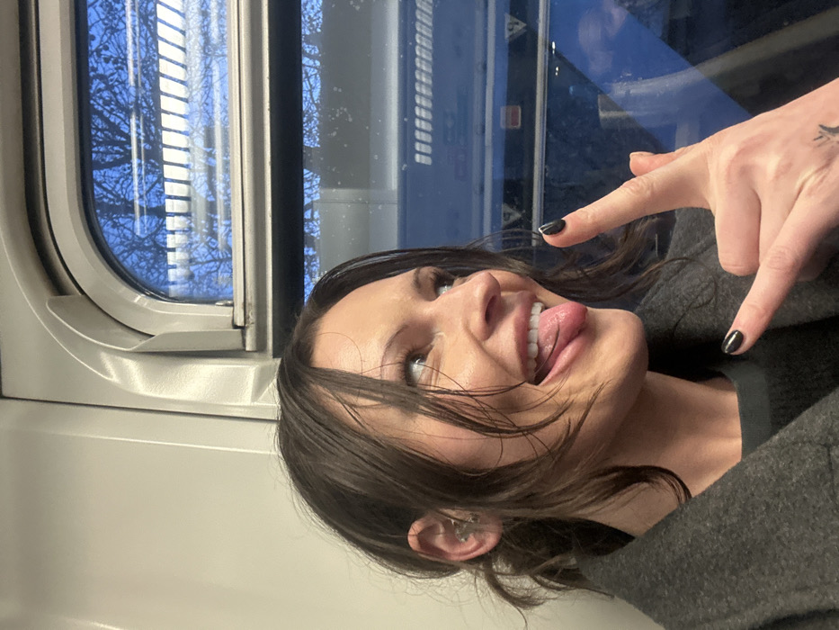 A woman with dark hair is sticking her tongue out and making the "rock on" hand gesture. She is on a train, as evidenced by the window showing trees outside. She has dark nail polish and a small tattoo on her hand. Her skin is light-toned. She wears a dark jacket.