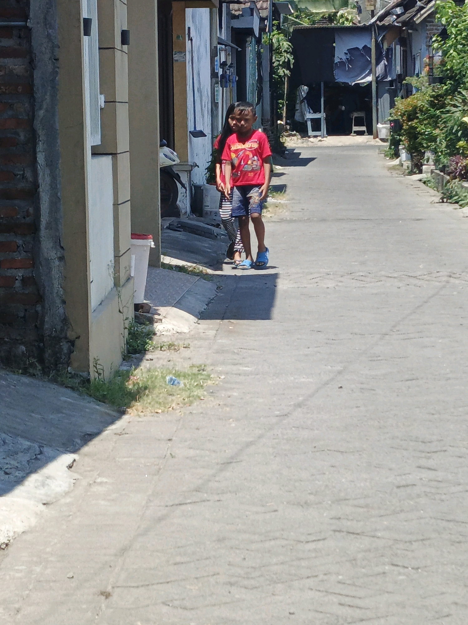 In the city of Ketegan, Indonesia, a child wearing a red t-shirt with a graphic print and blue shorts walks down a paved street. The child is wearing blue sandals. To the left of the child, another person is partially visible, wearing a black and white striped shirt. The street is lined with buildings and vegetation. The sun is shining, casting shadows on the ground. The scene appears to be a residential area in Indonesia.