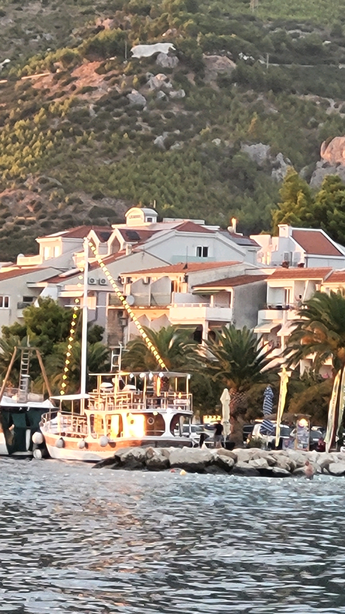 In Općina Tučepi, Croatia, a body of water with rippling surface reflects the warm, golden light of what appears to be late afternoon or early evening. A rocky shoreline borders the water, with a moored boat prominently featured. The boat is illuminated by strings of lights, and its deck is visible, showing some seating and a few people. Behind the boat, a cluster of white buildings with red-tiled roofs is situated on a hillside covered in dense green vegetation. The hillside rises steeply behind the buildings, with rocky outcrops visible. The scene depicts a coastal town setting during a pleasant time of day.