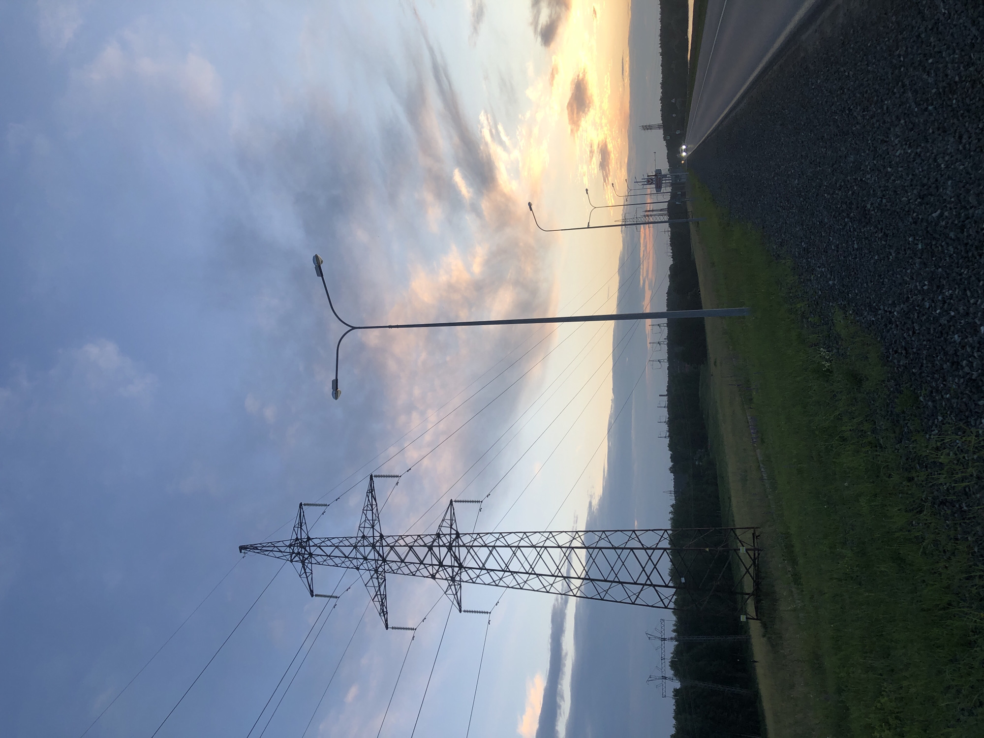 The image shows a sunset over a rural landscape with a road, power lines, and streetlights. The sky is filled with clouds tinged with orange and yellow hues. On the left side of the image, a tall electrical pylon stands prominently, with several power lines stretching across the frame. A grassy field and a line of trees are visible behind the pylon. To the right, a paved road runs alongside a gravel shoulder. A series of streetlights line the road, with the furthest ones appearing to illuminate oncoming car headlights. The scene suggests an evening view on the outskirts of Chaikovsky, Russia, possibly near a major thoroughfare given the presence of streetlights and the road. No people are visible in the image.