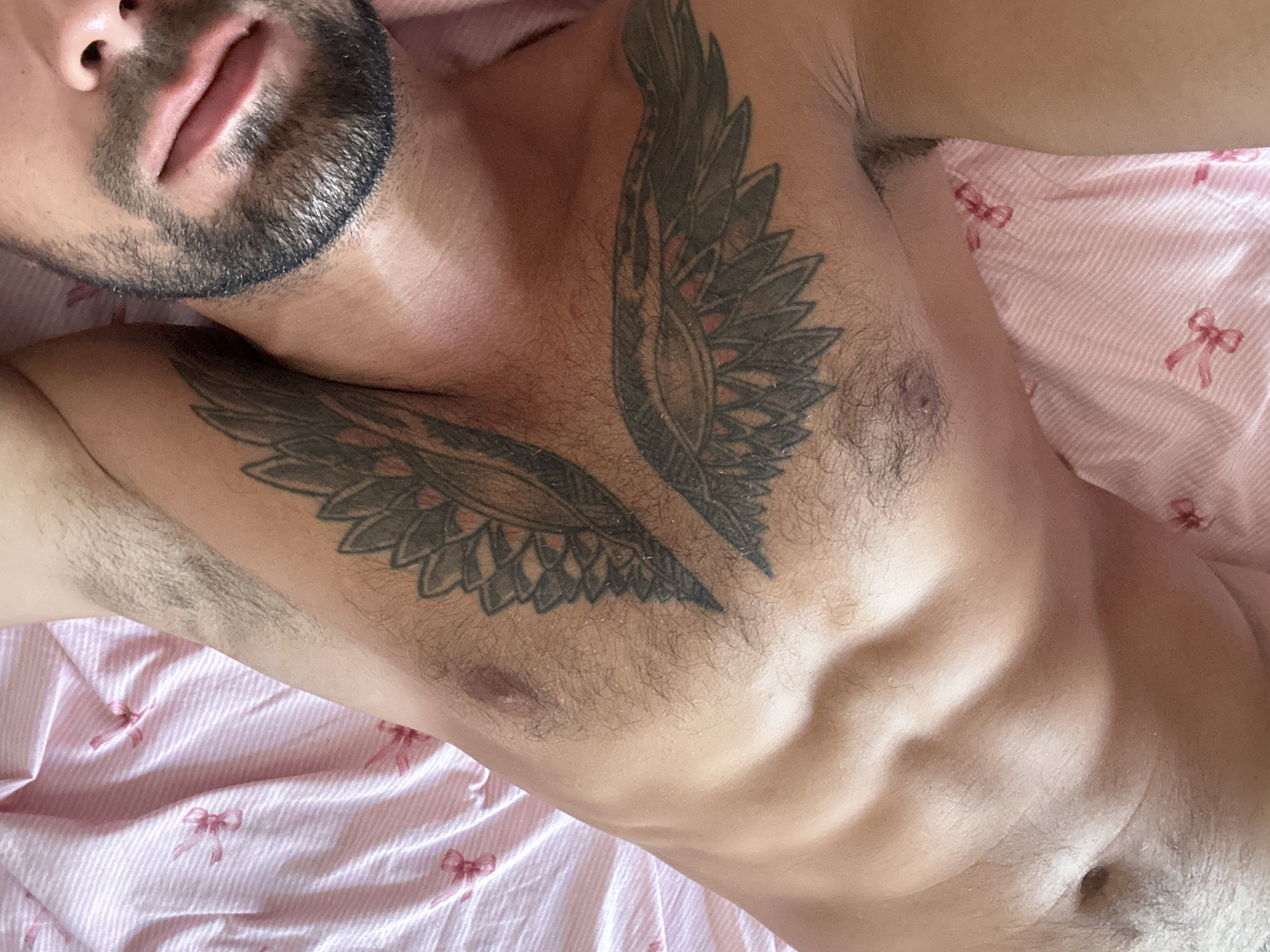 The image shows a close-up, overhead view of a person's torso lying on a pink, striped sheet with small bow patterns. The person has dark hair on their chest and arms, and a beard. Two wing tattoos are visible on their chest and upper arm. The person's mouth and part of their face are visible in the upper right portion of the image. The scene appears to be indoors, possibly a bedroom, with no discernible elements of the city of Nha Trang Ward, Vietnam.