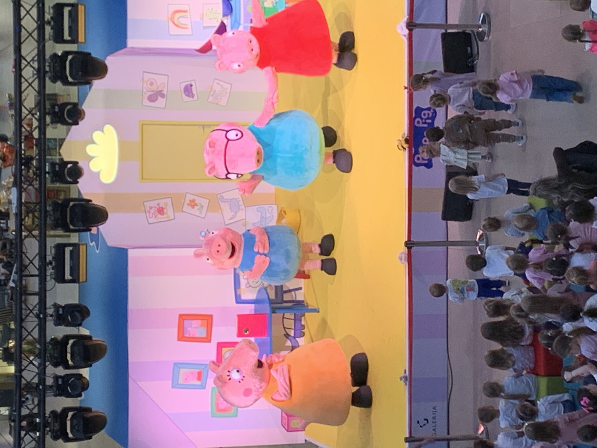 This image captures a lively indoor event, likely a stage show or meet-and-greet, featuring characters from the "Peppa Pig" animated series. Four large costumed characters are prominently displayed on a brightly lit stage: Peppa Pig in a red dress, George Pig in a blue top and glasses, Mummy Pig in a light blue dress, and Daddy Pig in an orange top. The stage is set with a vibrant yellow floor and a whimsical backdrop featuring a sky-blue upper section and a pink-striped lower wall decorated with children's drawings, including a yellow cloud shape, butterflies, and various framed pictures of Peppa Pig scenes.
An audience, primarily composed of young children and accompanying adults, is gathered to the right of the stage. Many children are seated on colorful blocks in the front rows, attentively watching the performance, while others stand further back. The atmosphere appears to be one of engagement and excitement, typical of a children's entertainment event. Stage lighting equipment, including several spotlights, is mounted on a metal truss structure visible on the far left, illuminating the performance area.
The event is taking place indoors, likely during the day, given the nature of the audience. Visible text includes "Peppa Pig" on a blue sign positioned near the audience section, clearly indicating the theme. Additionally, the word "GALERIJA" is visible on a grey vertical panel to the far right, accompanied by a small, abstract logo. This suggests the event is held at "Galerija," potentially referring to Galerija Belgrade, aligning with the provided location context of Belgrade, Serbia. The characters' poses imply they are actively performing or interacting with their audience.
