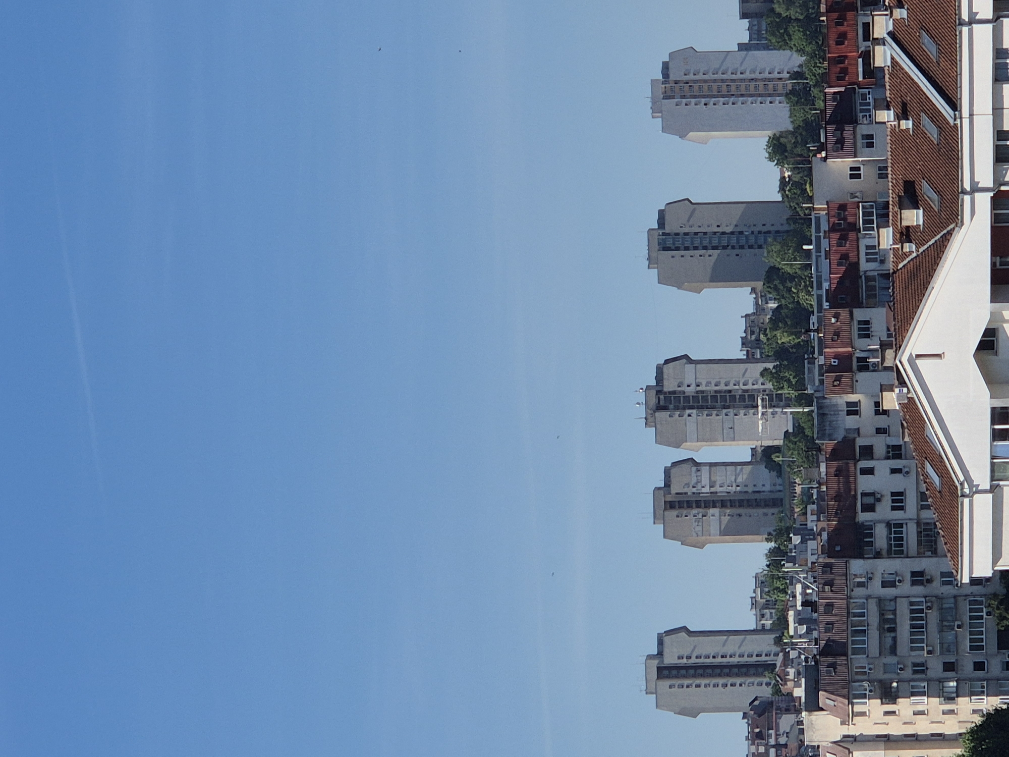 A medium shot captures a cityscape under a clear blue sky. In the foreground, several multi-story buildings with red-tiled roofs are visible. Behind them, a row of taller, grey apartment buildings stands out against the sky. Five identical, distinctive grey towers with protruding upper sections are spaced evenly in the middle ground, appearing to be part of a residential complex. The sky is clear with a faint contrail visible in the upper left quadrant. The overall impression is one of urban development with a focus on apartment living.