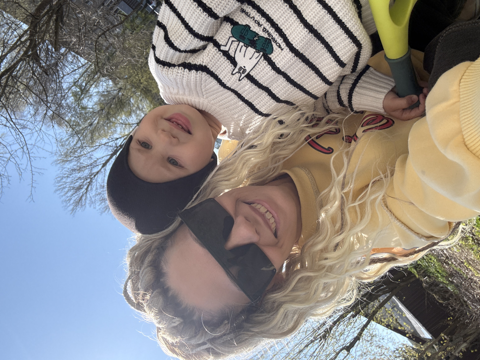 The image shows a selfie taken from a low angle looking up at a woman and a young child. The woman has long blonde hair, is wearing sunglasses, and is smiling. She is wearing a yellow sweatshirt. The child is wearing a black beanie and a striped white and black sweater with a skateboard graphic. The child is also smiling. They appear to be outside on a sunny day, with bare trees visible in the background. The location is suggested to be Rusanovo, Russia.