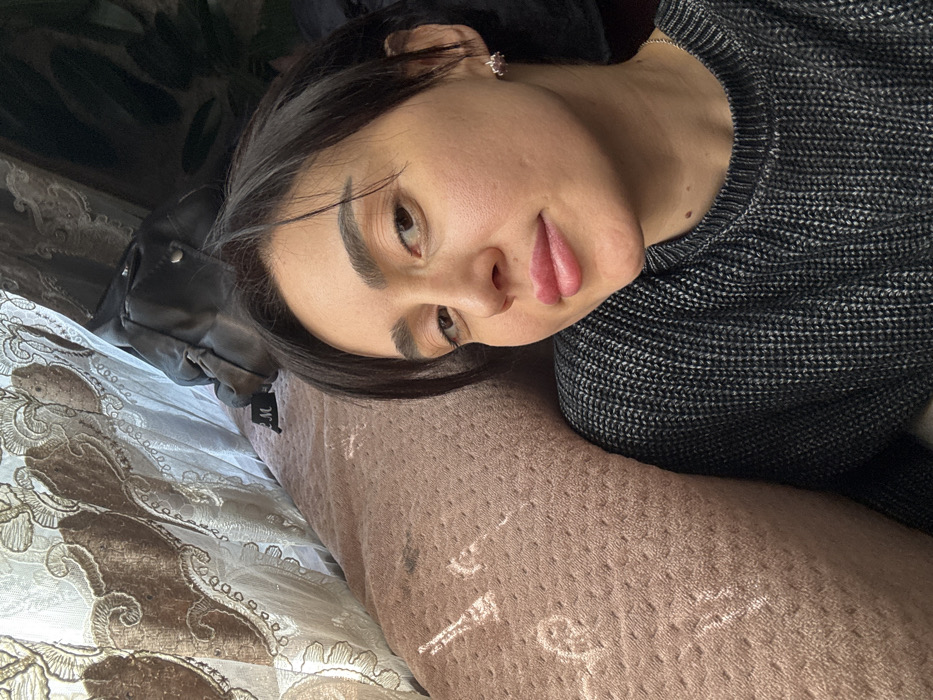 The image depicts a close-up indoor portrait of an adult female, likely situated in a residence in Bolnisi, Georgia. The subject is a woman with short, dark hair, light-colored skin, and full lips, wearing a dark grey knitted sweater and a small stud earring in her visible right ear. She is angled slightly, looking directly at the camera with a subtle smile. Her head rests against a textured, light brown or pinkish fabric surface, possibly a pillow or blanket, which shows minor dark smudges and lighter marks. In the background to the upper left, an ornate, translucent light-colored curtain with brown or gold embroidered patterns is visible, through which light appears to be filtering, casting subtle illuminated patterns on the adjacent fabric surface. To the upper right, behind the subject's head, a dark object resembling a black leather jacket or bag is partially visible, along with a small black tag marked with white lettering (possibly "M" or "EM"). No other individuals or specific actions are depicted.