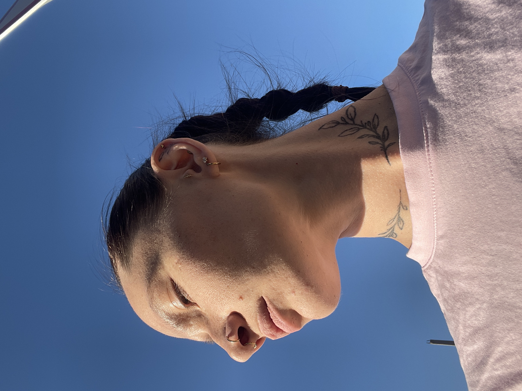 A woman is seen from a low angle, with a clear blue sky in the background. The woman has dark hair, styled in a braid, and has several piercings in her ear. She also has a nose ring. The woman's skin is fair, and she is wearing a light pink top. A tattoo of a plant is visible on her neck. The scene appears to be outdoors, likely in a sunny area. There is no indication that the photo was taken in Pervomaysky, Russia.