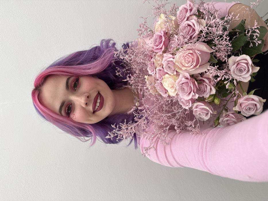 A female individual is shown from the mid-chest up, centered against a plain white background. She possesses wavy, bi-colored hair, specifically pink at the crown and front, fading to purple along the sides. Her facial expression is a wide smile, revealing dimples. Makeup includes prominent pink eyeshadow, winged eyeliner, dark red lipstick, and a small red heart drawn on the left cheekbone. She wears a white beaded necklace and a light pink, long-sleeved, ribbed top. The individual holds a substantial bouquet of flowers with both hands. The bouquet is composed of light purple, light pink, and cream-colored roses, accented with reddish-pink dried or preserved branched filler foliage. The scene is located in the City of Zagreb, Croatia.