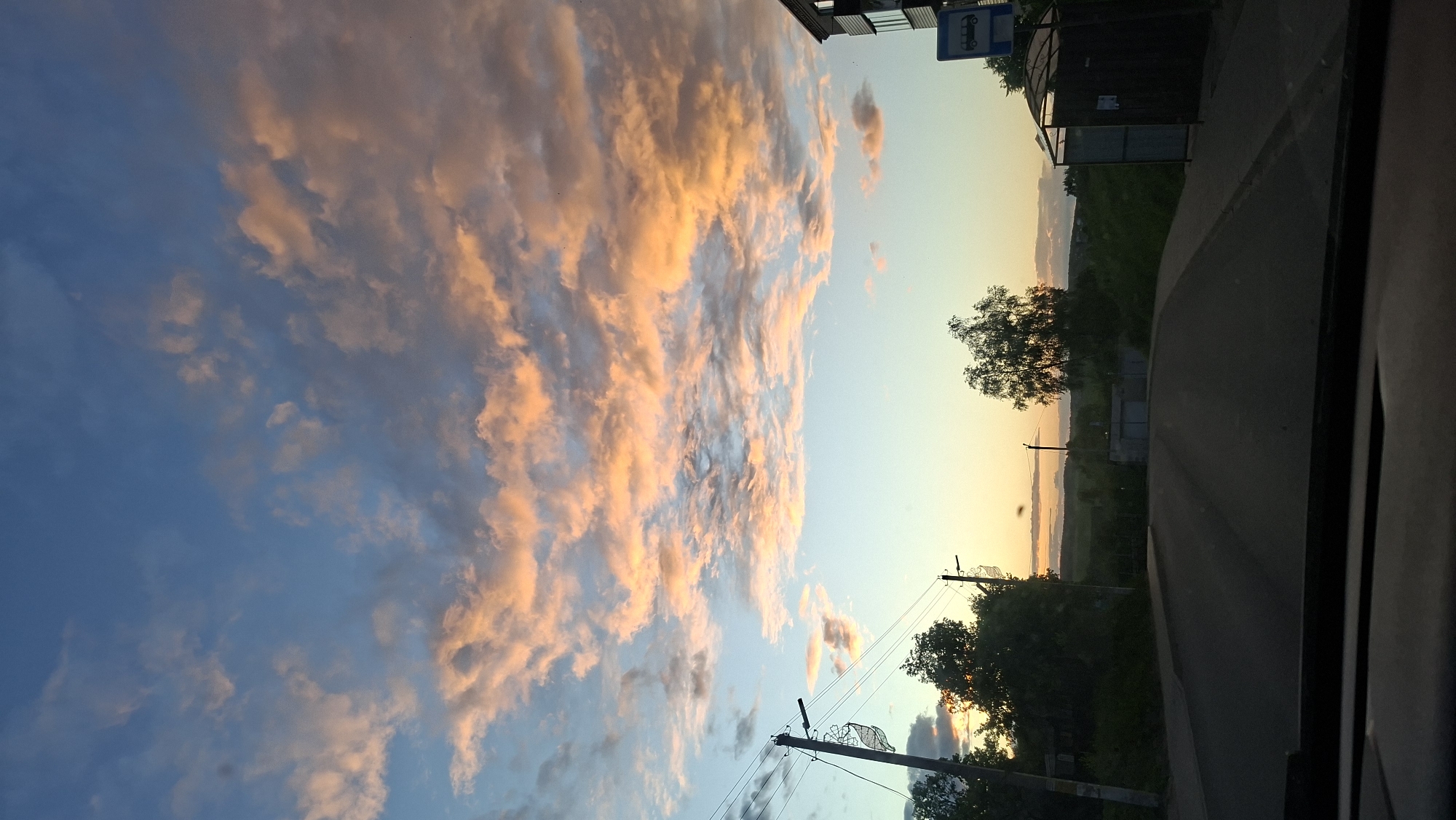 The image captures a scene in or near Смалявічы, Belarus, at dusk or dawn. The sky is predominantly blue with a dramatic display of orange-tinged clouds. A road runs through the right side of the frame, with green vegetation and trees lining it. Utility poles and wires are visible near the road. In the upper right corner, what appears to be a bus stop structure is present. The shot is taken from inside a vehicle, as indicated by the car window frame on the right. The overall impression is of a rural or suburban environment at a time when the sky is illuminated by the setting or rising sun.