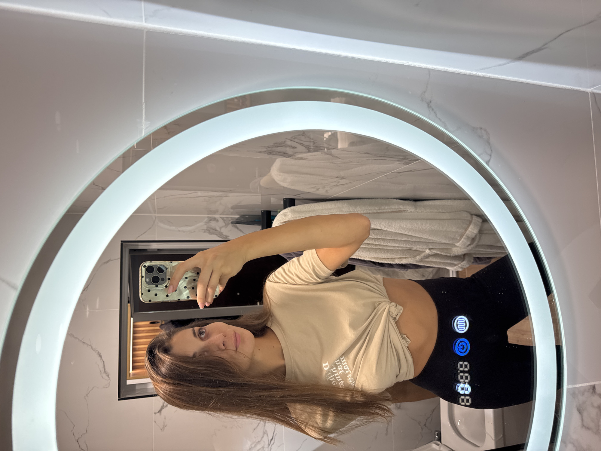 A young woman is taking a selfie in a bathroom mirror. The mirror is round with a light ring around it. The woman is wearing a tan T-shirt tied at the waist and black leggings. The leggings have some electronic buttons and a digital clock display, which are integrated into the garment. The bathroom has marble-patterned tiles. The location is likely Burgas, Bulgaria.