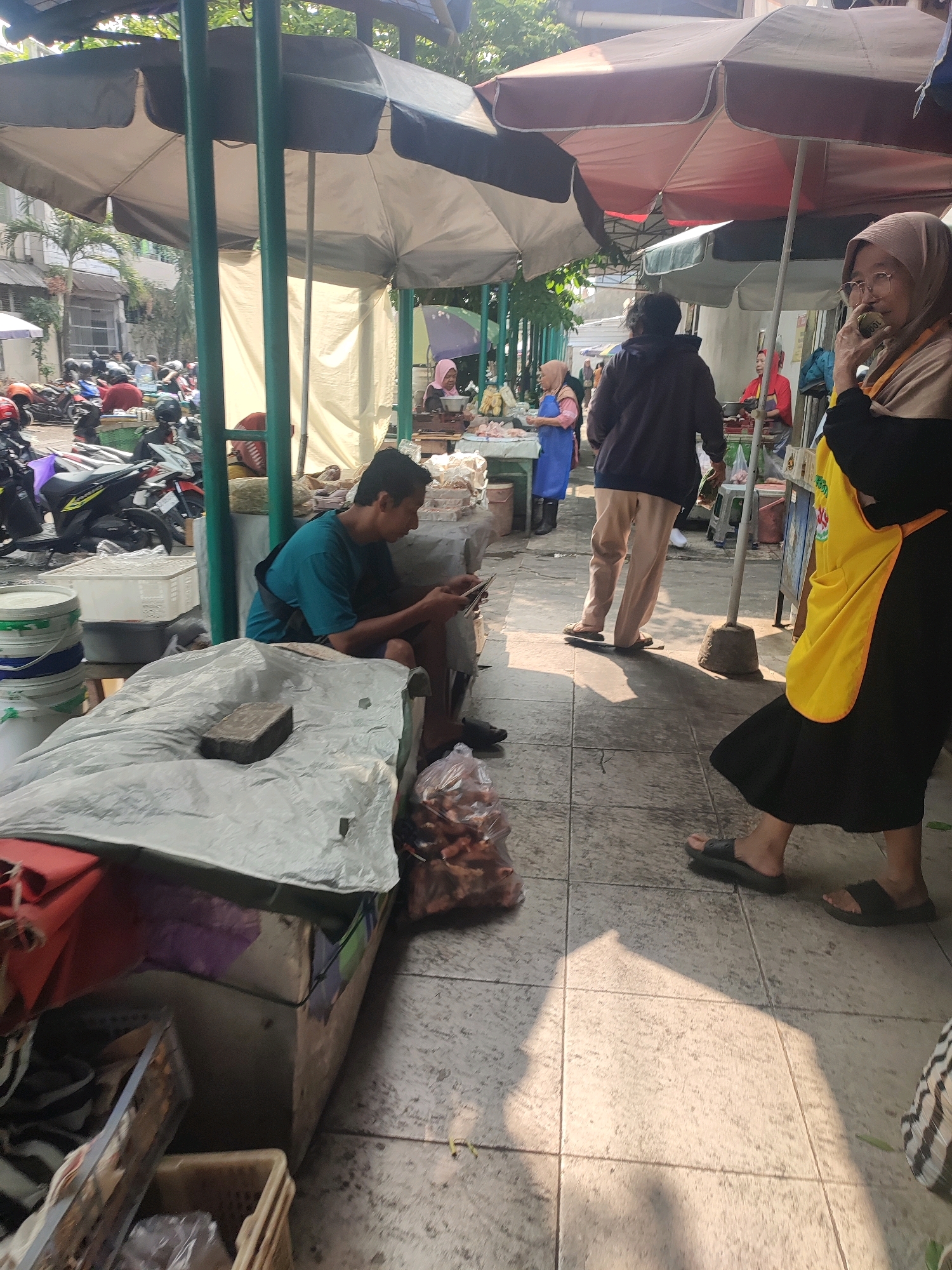 The image depicts an outdoor market scene in Yogyakarta, Indonesia, likely a street-side market or a traditional market on a bright, sunny day. The ground is paved with square tiles, and there is a strong presence of sunlight and shadows, suggesting it is daytime.
In the foreground, a young man wearing a teal t-shirt is seated and focused on his mobile phone. He is positioned behind a makeshift table covered with a grey tarp, on which a brick and some packaged goods are visible. To his left, there are stacks of buckets, one with a blue and white rim.
Further into the scene, there are several stalls or vendors under large umbrellas. One stall appears to be selling food items, with bags of what looks like dried goods or snacks. A woman in a hijab and a yellow apron is standing near a stall, holding a small object to her mouth, possibly eating or talking on a phone. She is wearing glasses and a dark dress. Another person, a woman in a blue apron, is visible behind a display of goods.
In the background, there are numerous motorcycles parked, indicating a bustling area with people commuting or shopping. Trees and buildings are also visible in the distance, framing the market setting. The overall atmosphere appears to be that of a typical Indonesian market, with vendors and customers interacting amidst various goods and parked vehicles.