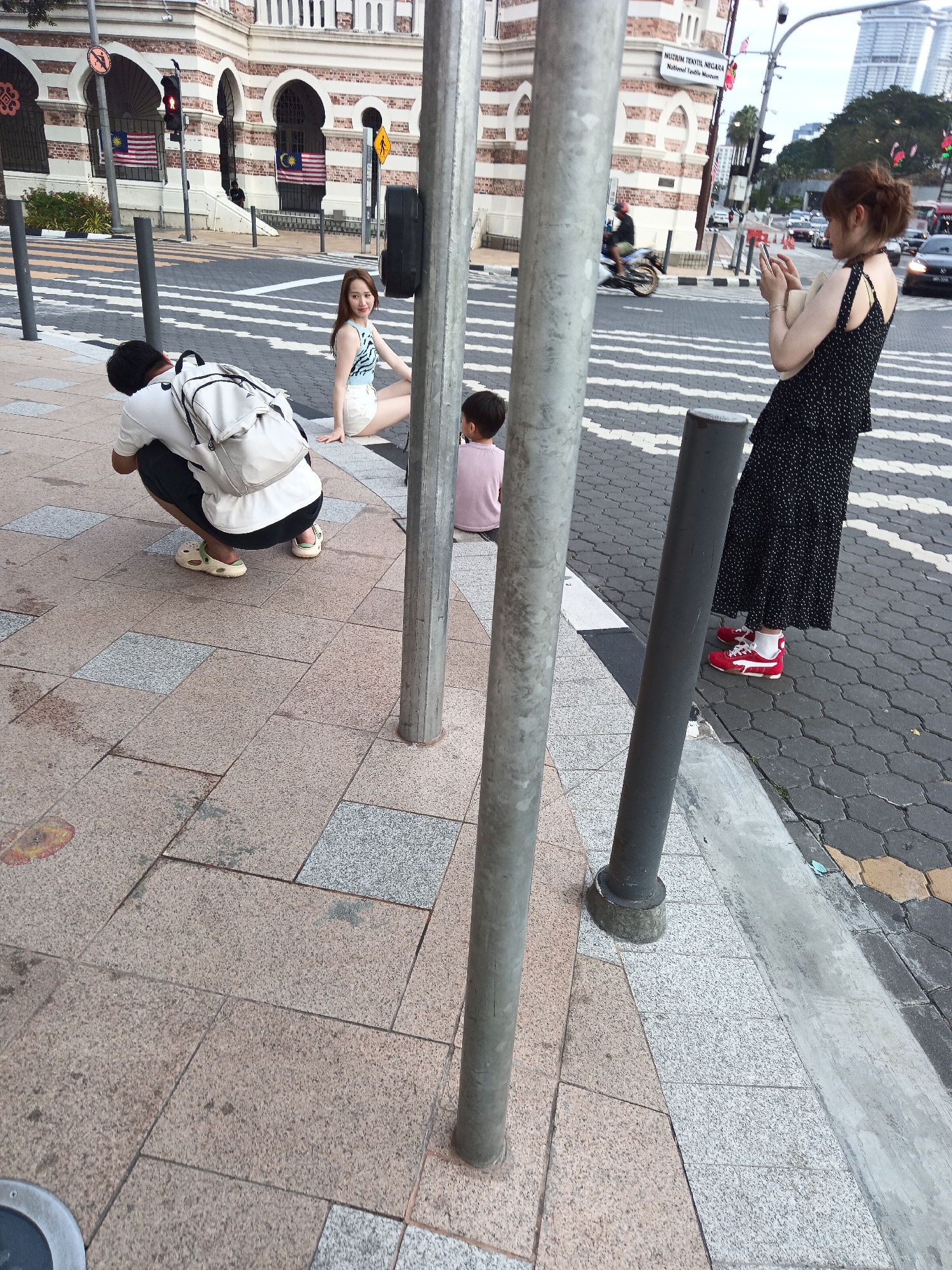 At the historic Independence Square in Kuala Lumpur, Malaysia, a group of individuals are captured in a moment of leisurely activity. A man in a white t-shirt and dark shorts, wearing a white backpack, is crouched down, seemingly taking a photograph of a young woman. The woman, dressed in a blue and white striped top and white shorts, is seated on the ground, looking towards the man. Beside her, a child in a pink t-shirt sits with their back to the camera. In the background, a woman in a black polka dot dress is standing and holding a phone, likely capturing her own memories of the day. The iconic Sultan Abdul Samad Building, with its distinctive Moorish architecture, stands proudly in the backdrop, while a busy street with traffic can be seen in the distance. The scene is set on a paved sidewalk, with a pedestrian crossing prominently featured.