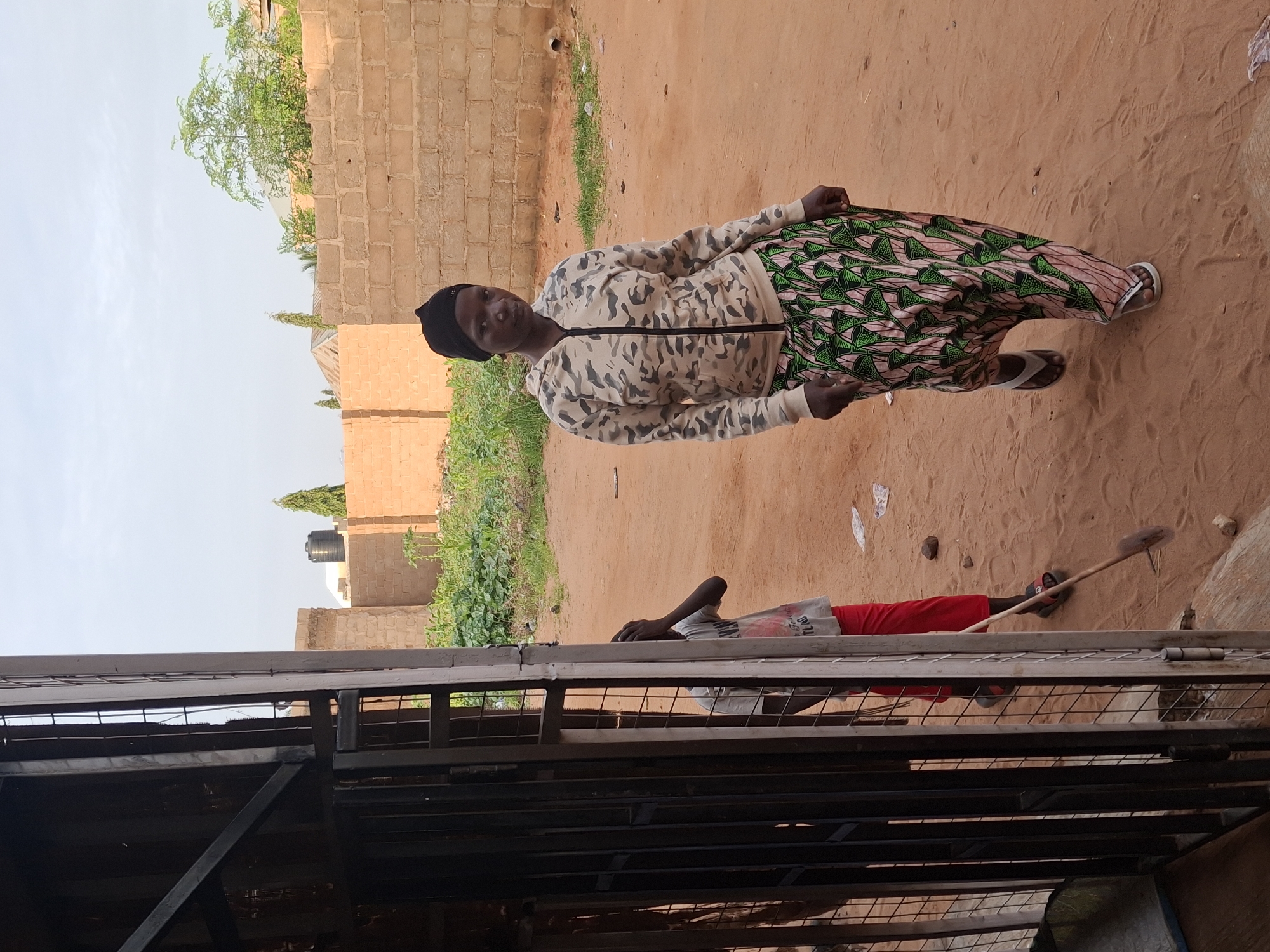 In Tumfure, Nigeria, an outdoor scene unfolds. A woman with dark skin and wearing a black headscarf, a camouflage zip-up hoodie, and a long, patterned skirt with green and pink designs is walking towards the camera. She has on white flip-flops. Beside her, to the left and partially obscured by a metal gate and fence, a young person wearing a light-colored t-shirt and red pants is visible. The child appears to be holding a shovel or rake. The ground is sandy and reddish-brown, with some sparse green vegetation and scattered debris. In the background, there is a brick wall made of light brown bricks, and beyond that, some buildings and trees under a hazy sky. A large black water tank is also visible on top of one of the structures. The lighting suggests it is either early morning or late afternoon.