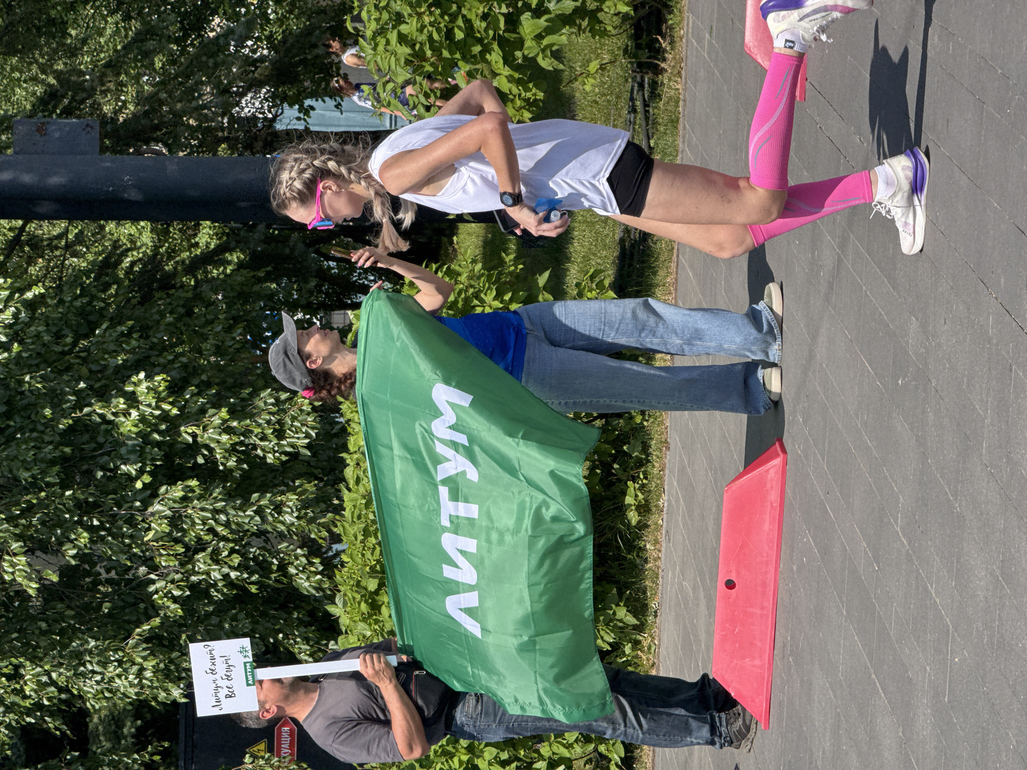 In Izhevsk, Russia, on a sunny day, a runner is seen passing a support crew holding a large green flag with the inscription "ЛИТУМ" in white letters. The runner, a woman with blonde braided hair and wearing pink compression sleeves and sunglasses, is in motion, her body angled towards the right of the frame. She is holding a water bottle in her right hand. To her left, a woman in a blue shirt and a baseball cap is also holding the flag, and to the far left, a man in a grey t-shirt and jeans holds a sign that reads "Лимум бежит? Все бегут!" (Is Litum running? Everyone is running!). The background features lush green trees and bushes, suggesting an outdoor urban environment, possibly a park or a street course during an event. A red, triangular barrier is visible on the pavement in the foreground.