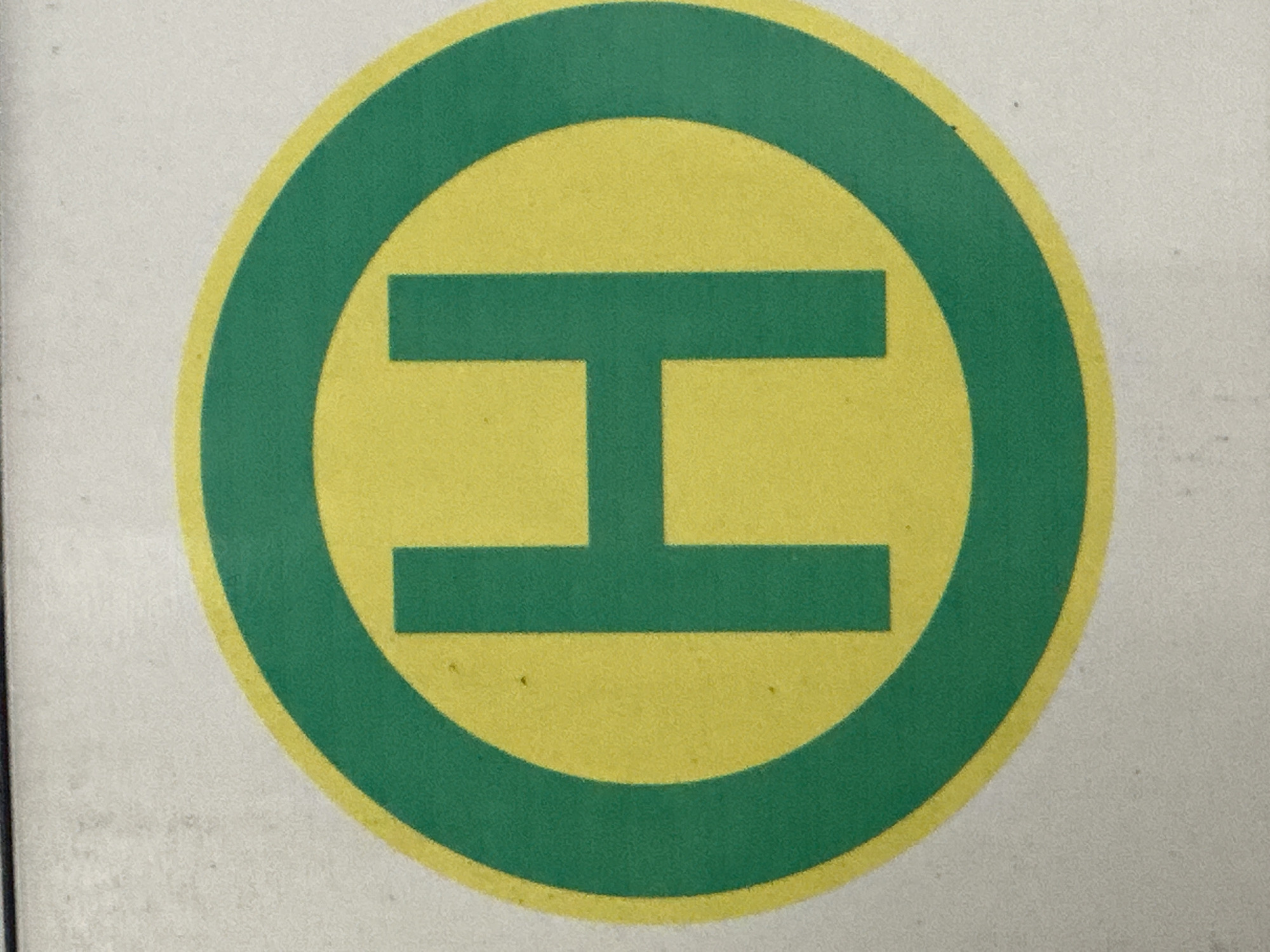 The image shows a close-up of a circular sign with a green border and a yellow center. In the center of the yellow circle is a large, bold, green letter "H". The sign appears to be a bus stop sign. The background is a light gray, textured surface, possibly a wall or a pole. There are no people or other objects visible in the image.