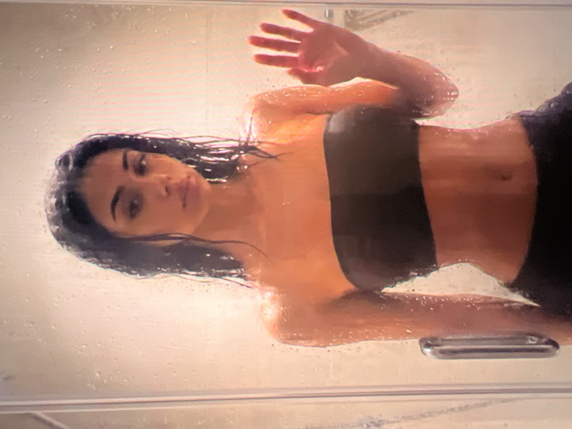 A young woman with dark hair stands in a shower stall, her body partially obscured by water droplets on the glass. She is wearing a dark strapless top that exposes her midriff. Her right hand is raised, with fingers spread, pressed against the shower glass. The background is indistinct and appears to be the interior of a shower.