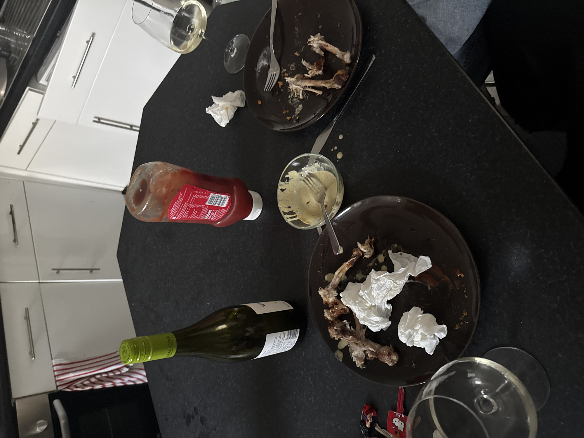 This image captures a close-up, overhead view of a dark kitchen countertop or table after a meal, likely in a domestic kitchen setting. The overall scene suggests a casual dining experience that has recently concluded.
The main subjects are various objects associated with a meal. Two dark brown plates are prominent, both containing remnants of food, specifically chicken bones (suggesting chicken wings or similar) and crumpled used paper napkins or tissues. One plate has a fork resting on it, while another fork is visible near the second plate. A small glass bowl holds a yellowish-white creamy sauce, with a fork dipped into it. Ketchup is present in a red plastic bottle with a white cap, bearing the text "KETCHUP" and "500ml".
For beverages, a partially full bottle of white wine, identified by its label as "CHÂTEAU COVE SAUVIGNON BLANC MARLBOROUGH NEW ZEALAND" (with "75cl e" and "12.5% VOL" also visible), lies on the table. Two wine glasses are also in view; one in the upper left holds a small amount of light-colored liquid (likely white wine), and another in the lower right is empty and resting on its side.
In the background, modern white kitchen cabinets with silver horizontal handles are visible, providing a context of a home kitchen. Other details include a red and white striped kitchen towel near the bottom left, and a set of keys with a "Hello Kitty" keychain near the bottom right. A person's leg or arm, clad in dark and light blue clothing, is partially visible in the upper right corner, indicating at least one person is still at the table. The lighting suggests an indoor setting, likely evening, given the lack of natural light.