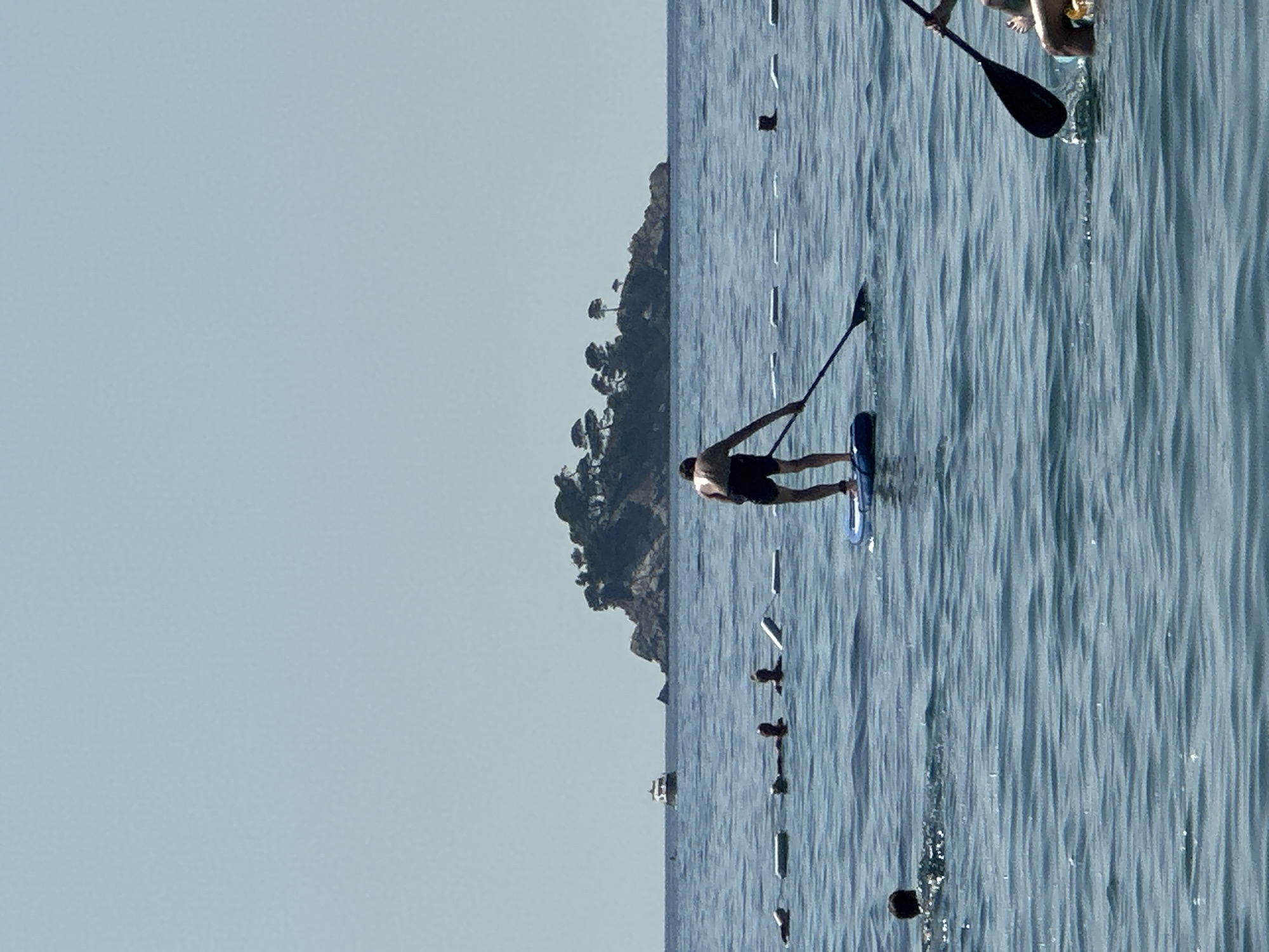 The image shows a body of water with a few people engaged in water activities. In the foreground, a person is paddleboarding and rowing with a paddle. To the left, other individuals are in the water, some with their heads visible. A floating barrier with buoys is present in the water, marking an area. In the background, a small island covered with trees and rocks is visible. A boat is also seen in the distance. The sky is clear and blue. The scene appears to be a beach or coastal area, likely in the vicinity of Đurovići, Montenegro, given the rocky island formation and clear water.