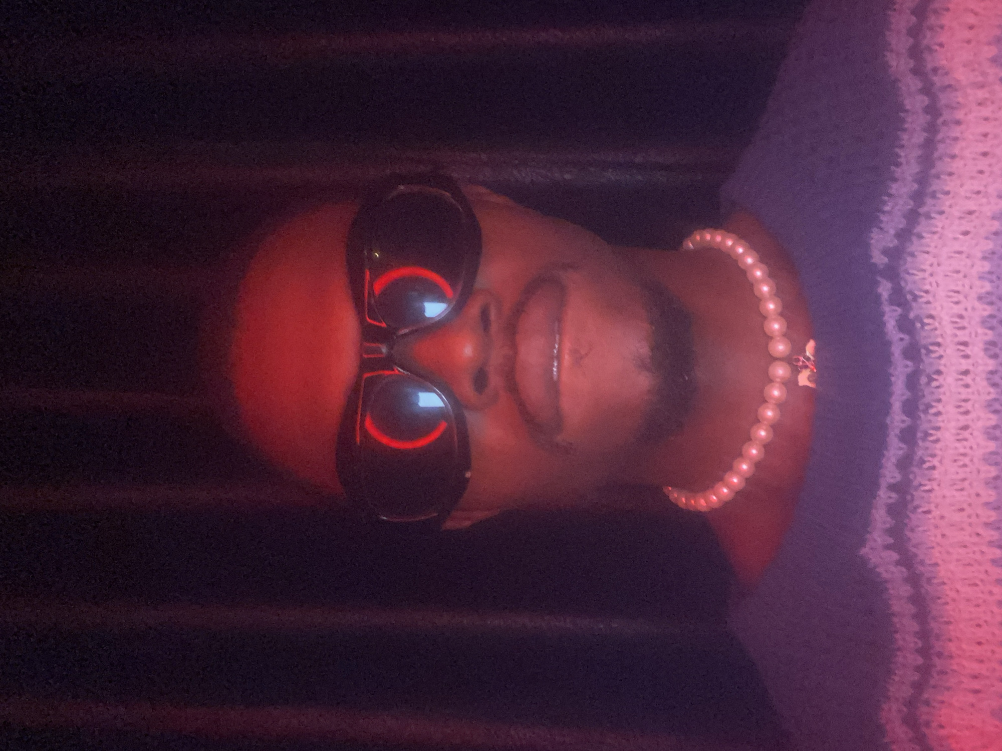 A person is shown from the chest up, facing the camera directly. They are wearing large, black, wraparound sunglasses that reflect a red, circular glow. A strand of pearl necklace is visible around their neck, with a small, light-colored charm hanging from it. They are also wearing a blue sweater with horizontal white stripes. The background is dark, with vertical lines suggesting a curtain or textured wall. The lighting is predominantly red, casting a warm, dramatic glow on the subject. The image appears to be a self-portrait taken in a dimly lit indoor setting. There is no information available about the location in Abakaliki, Nigeria.