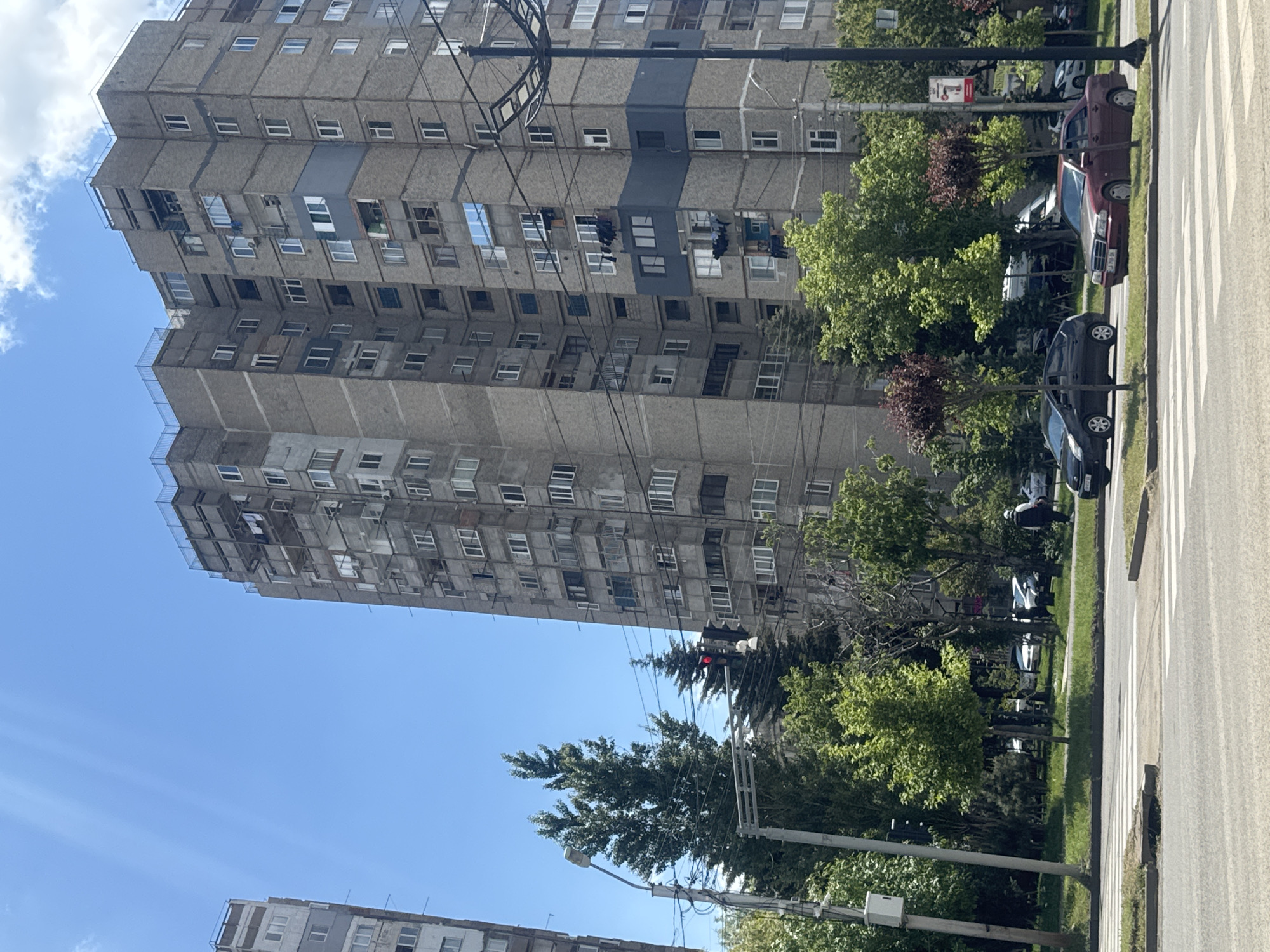 The image shows a high-rise apartment building in Tbilisi, Georgia. The building is concrete and has many windows. There are trees and cars on the street in front of the building. The sky is blue with some clouds. A traffic light is visible with a red light. A zebra crossing is visible on the road. A second building is visible in the bottom left of the image. Clothes hang from some of the windows of the building.