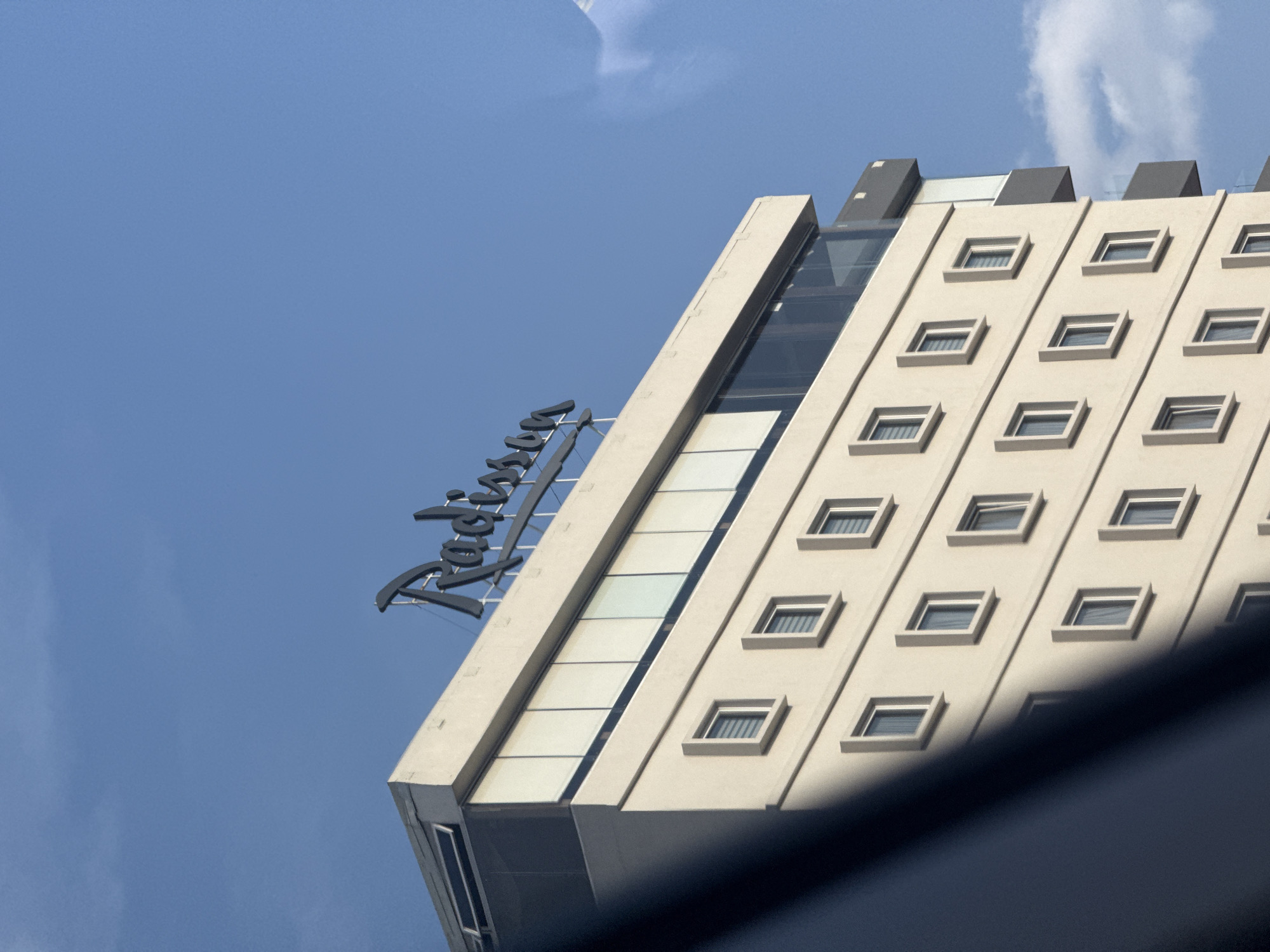 A low-angle, daytime shot showcases the upper corner of a Radisson hotel against a clear blue sky with a few scattered clouds. The hotel's facade is primarily a light beige color, with rows of square windows spaced evenly apart. The Radisson sign, in dark lettering, is affixed to the top of the building's edge. The hotel appears to be a modern, multi-story structure with clean lines and a minimalist design. The image was taken in Puebla, Mexico.