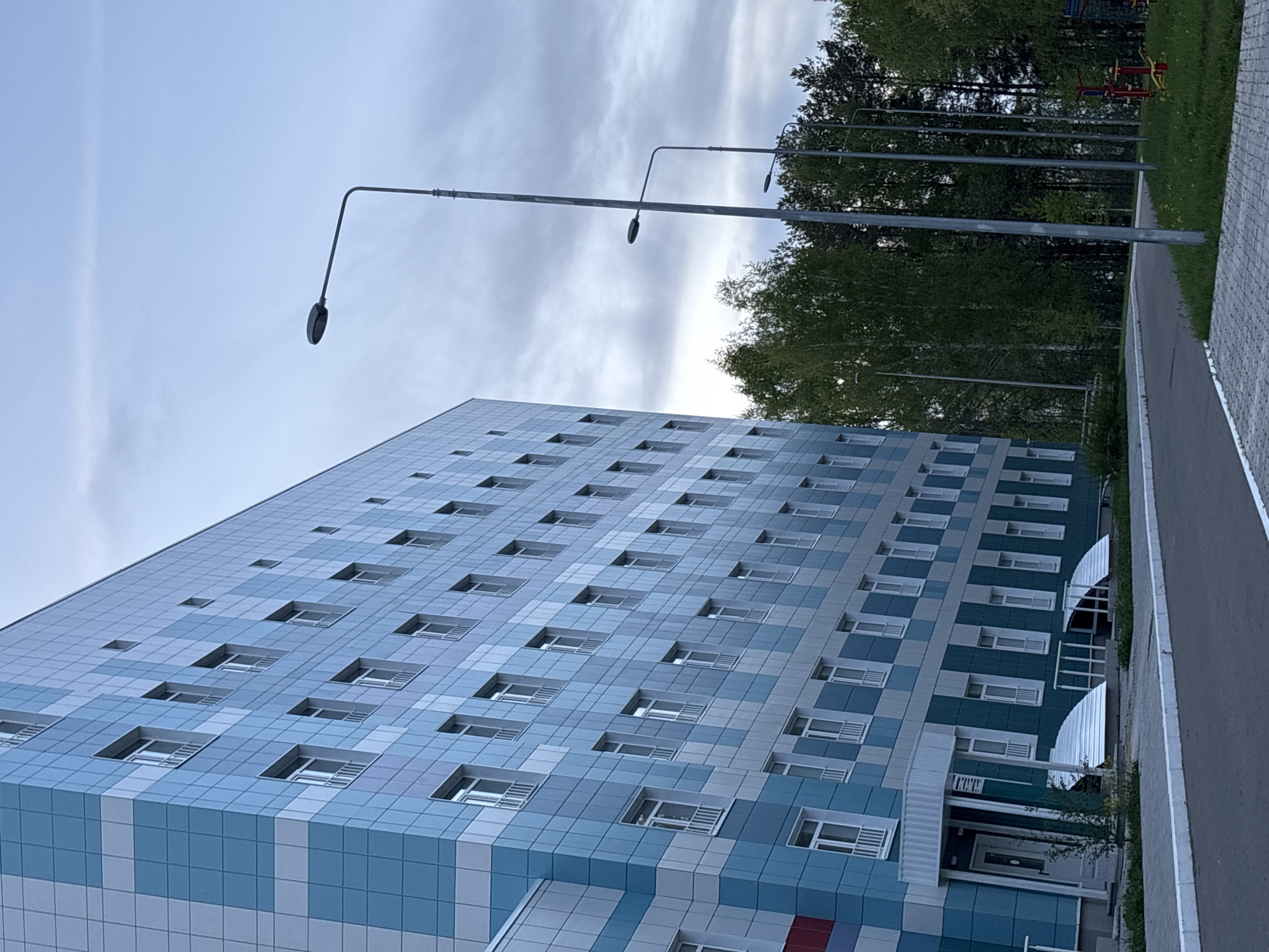 The image shows a tall, rectangular building with a grid-like façade of blue and white panels. The building has numerous square windows, appearing to be an apartment complex. The building is located in the city of Chaikovsky, Russia. Tall streetlights are visible in the background, along with trees and a paved pathway adjacent to the building. The sky is overcast with gray and white clouds. A small playground with red exercise equipment is visible in the background behind the row of trees.
